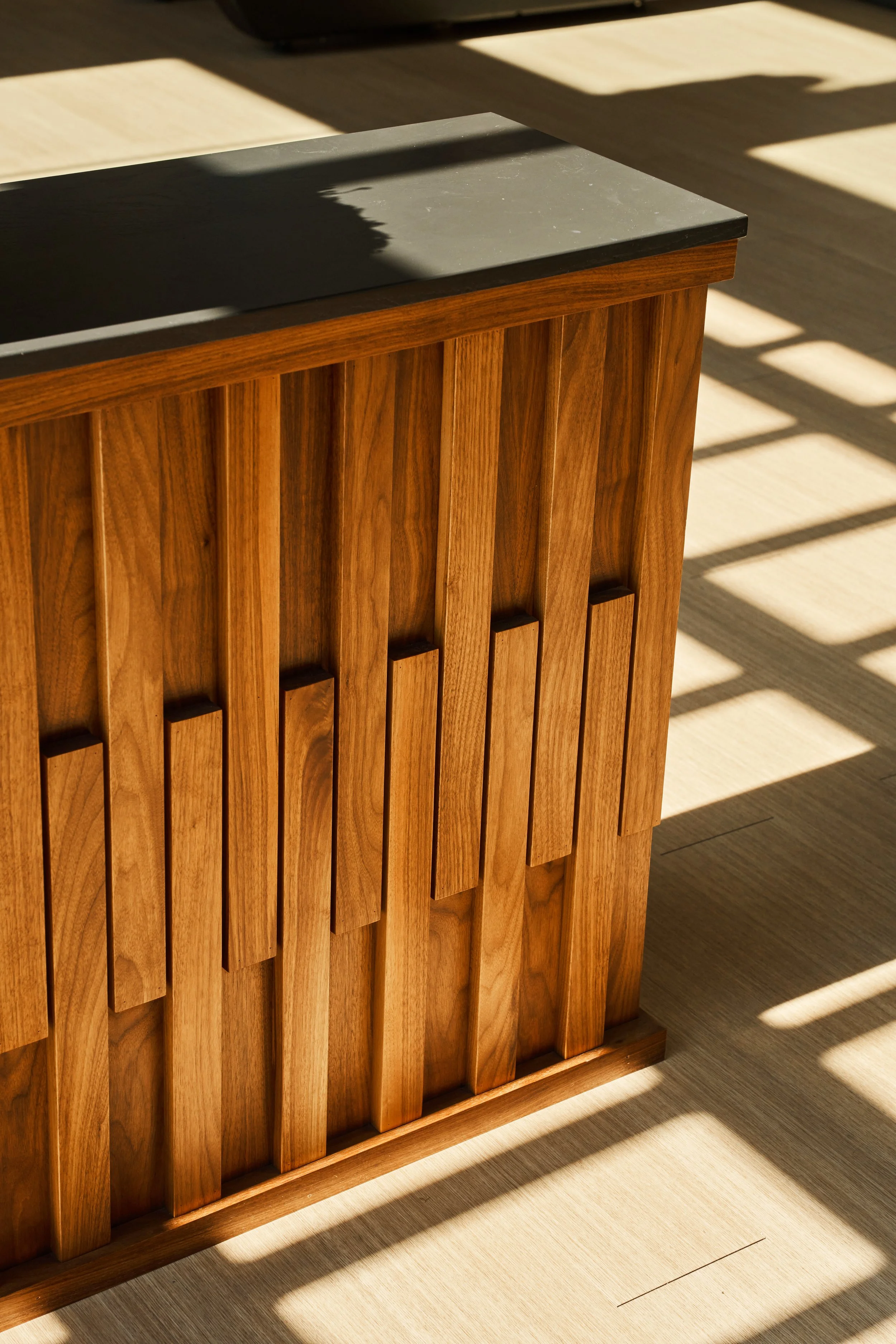 A wooden cabinet with vertical slats and a dark gray stone top, placed on a wooden floor with sunlight and shadows nearby.