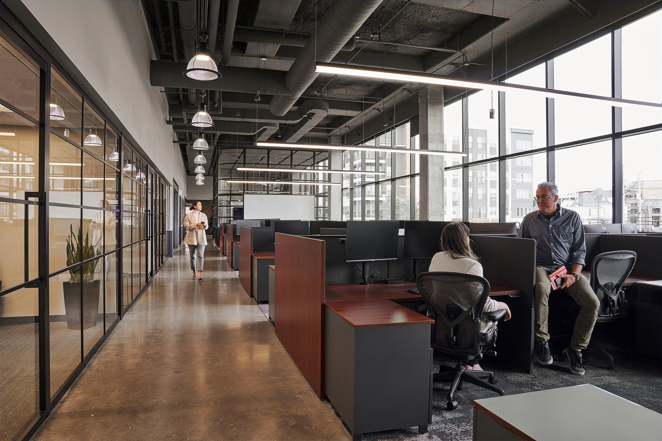 Modern office space with large windows, exposed ceiling pipes, and a few people working and walking. Open workstations and glass-partitioned rooms are visible.