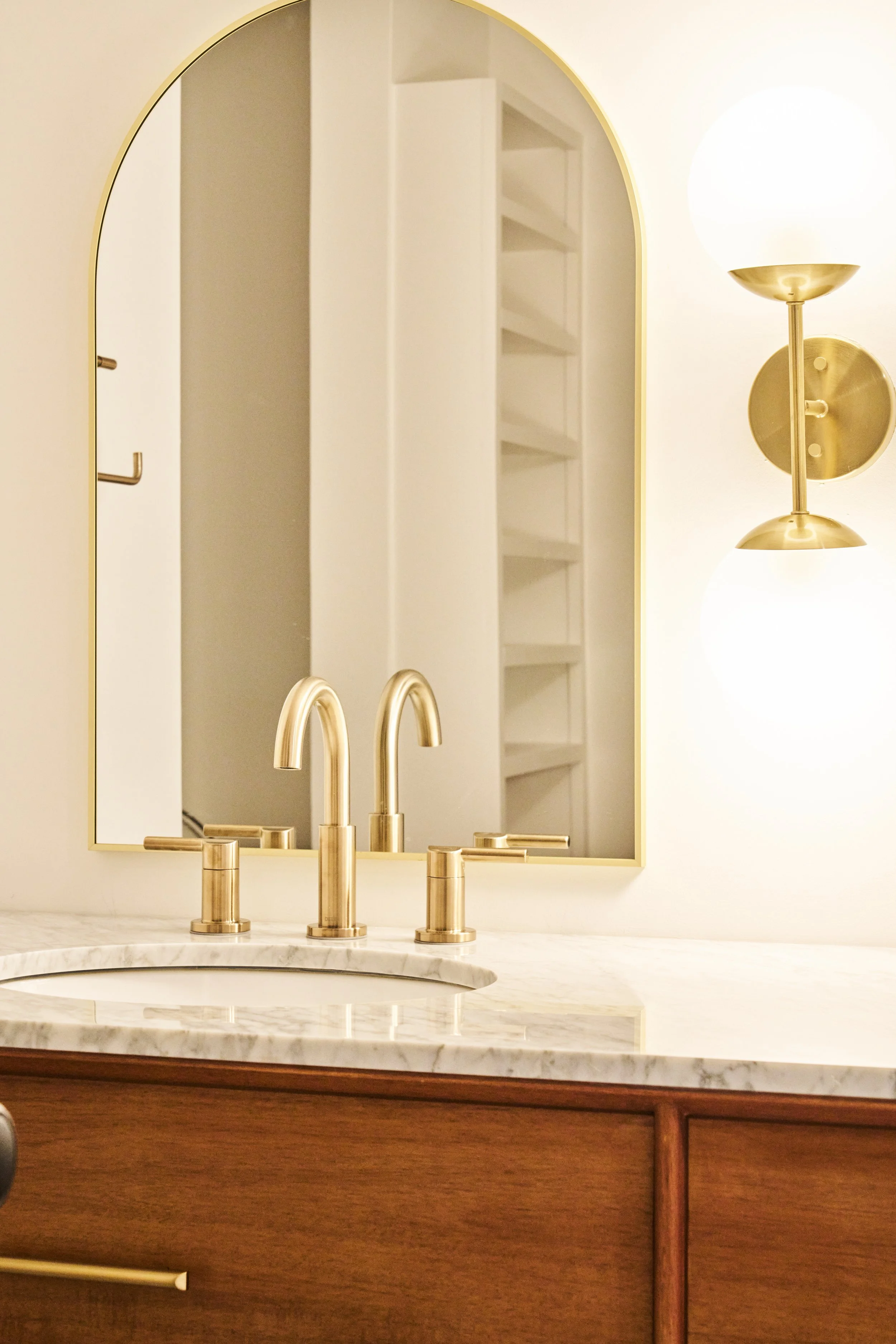 A bathroom vanity with a marble countertop, a gold-colored faucet, a mirror with a gold frame, a wall-mounted light with a gold base and white globe, and a white built-in shelving unit reflected in the mirror.