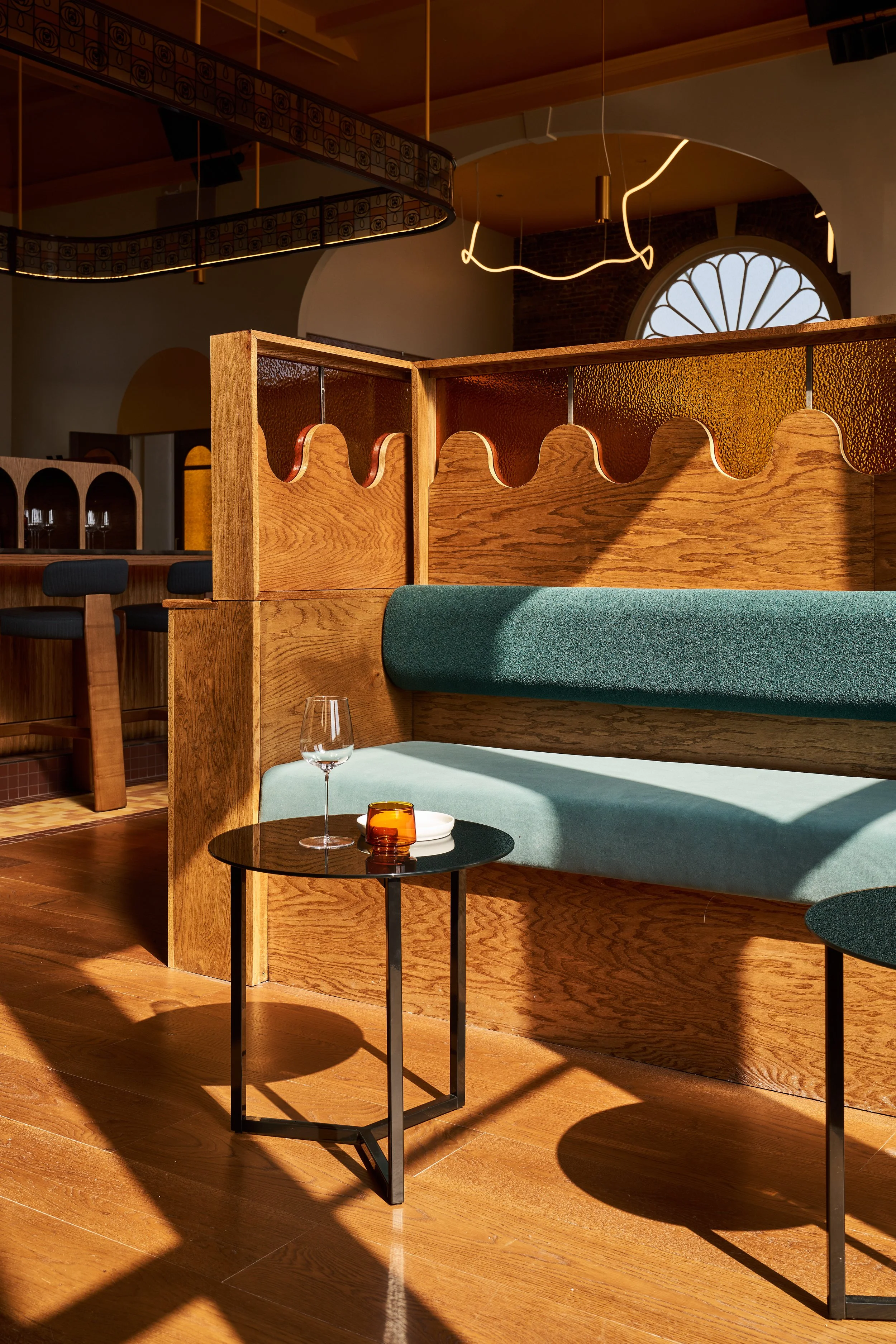 Interior of a modern restaurant with wooden accents, green upholstered seating, small round tables, and decorative lighting fixtures, including a large window with a rounded sunburst design.