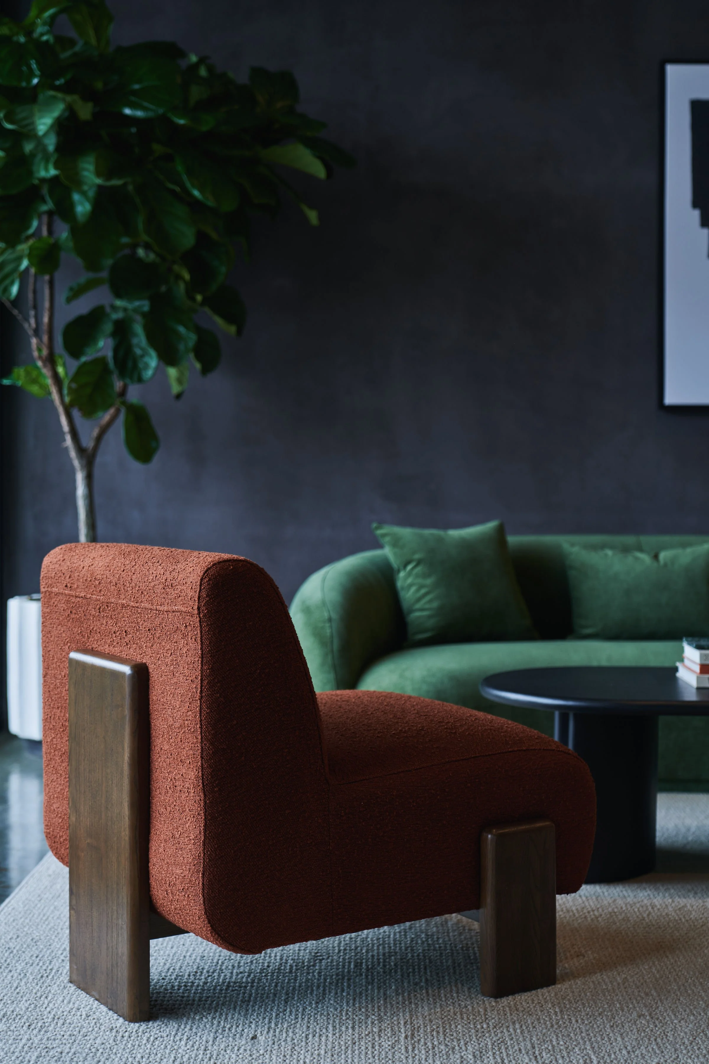 A reddish-brown upholstered armchair with wooden armrests and legs in a room with a gray carpet. Behind it, a green upholstered sofa with green cushions. To the left, a large green leafy plant. On the right, a black circular side table with books on 