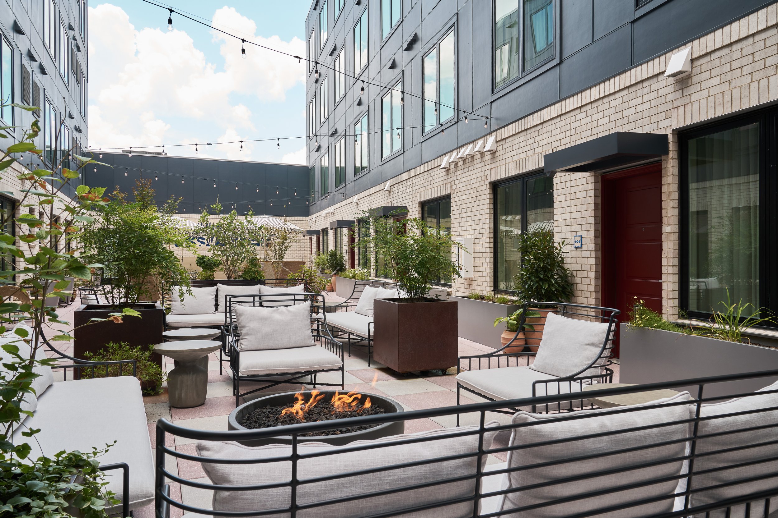 Outdoor patio with seating area, potted plants, string lights, and a fire pit surrounded by modern buildings with large windows.