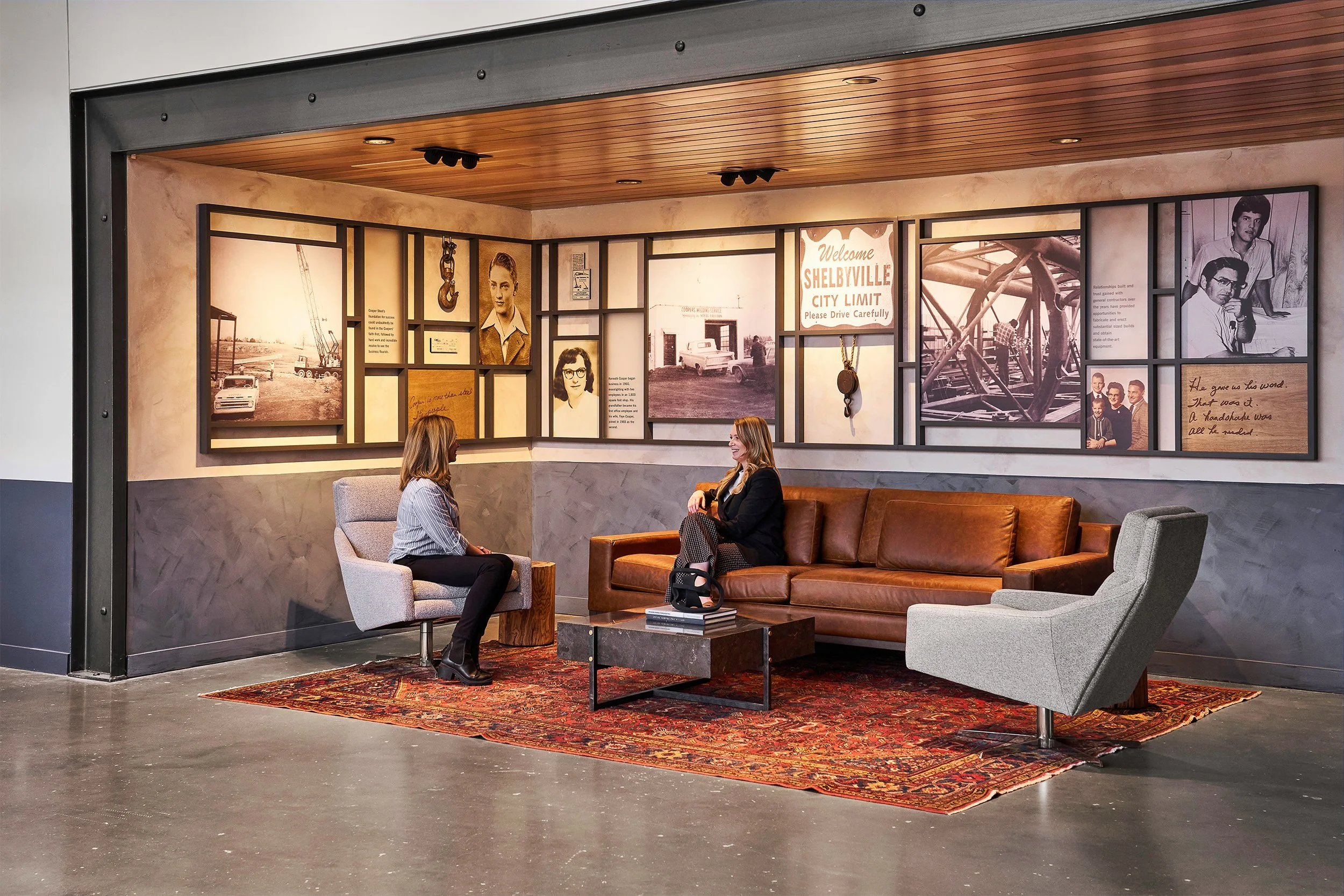 Two women sitting on modern chairs and a sofa in a lobby or waiting area with a collection of vintage photos and memorabilia on the wall behind them, including a sign that reads "Welcome Shelbyville City Limit" and a large red rug on the floor.