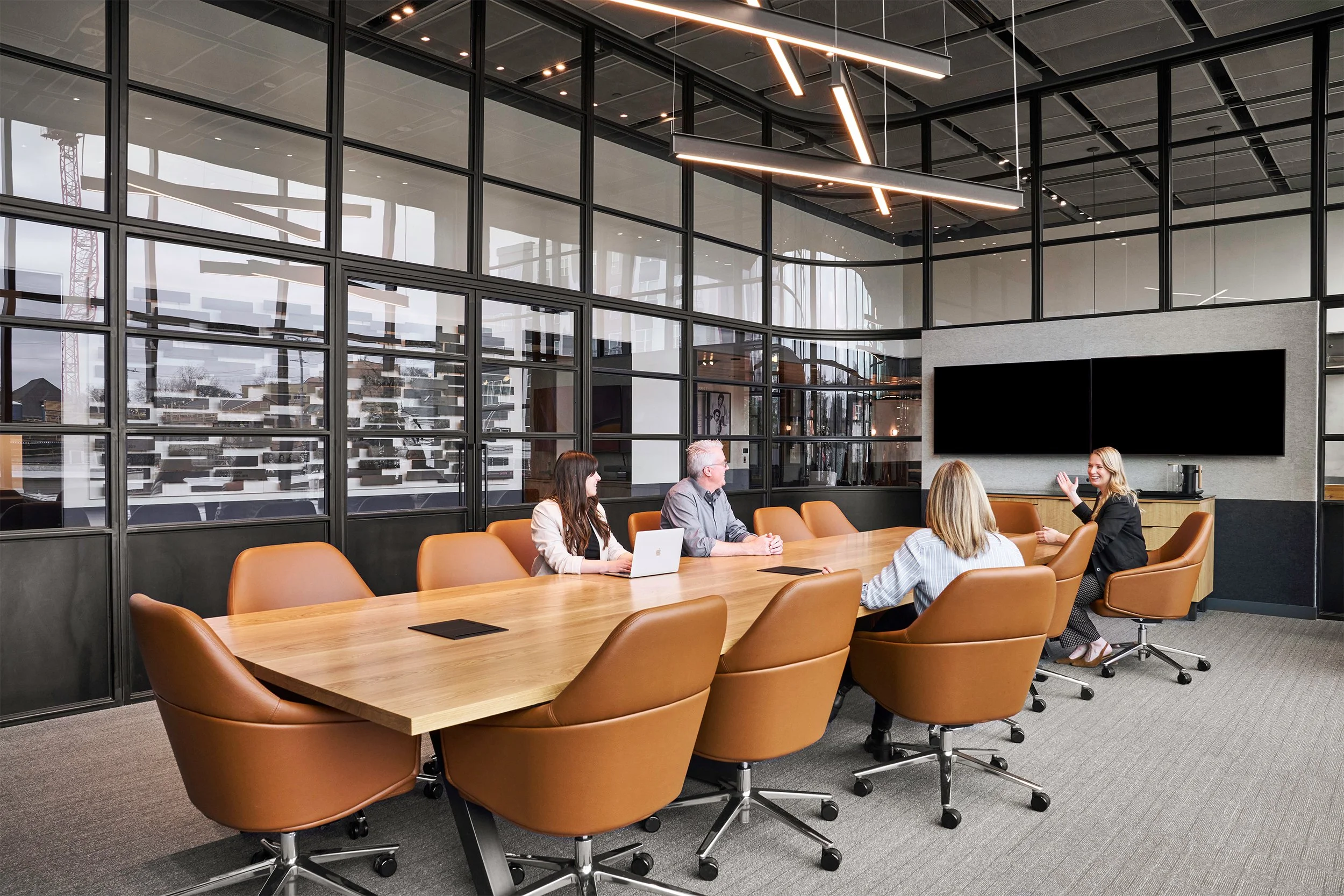 A business meeting taking place in a modern conference room with a large wooden table and brown leather chairs. Four people are engaged in discussion, with one woman speaking and three others listening. The room has a glass wall with a view of the ou