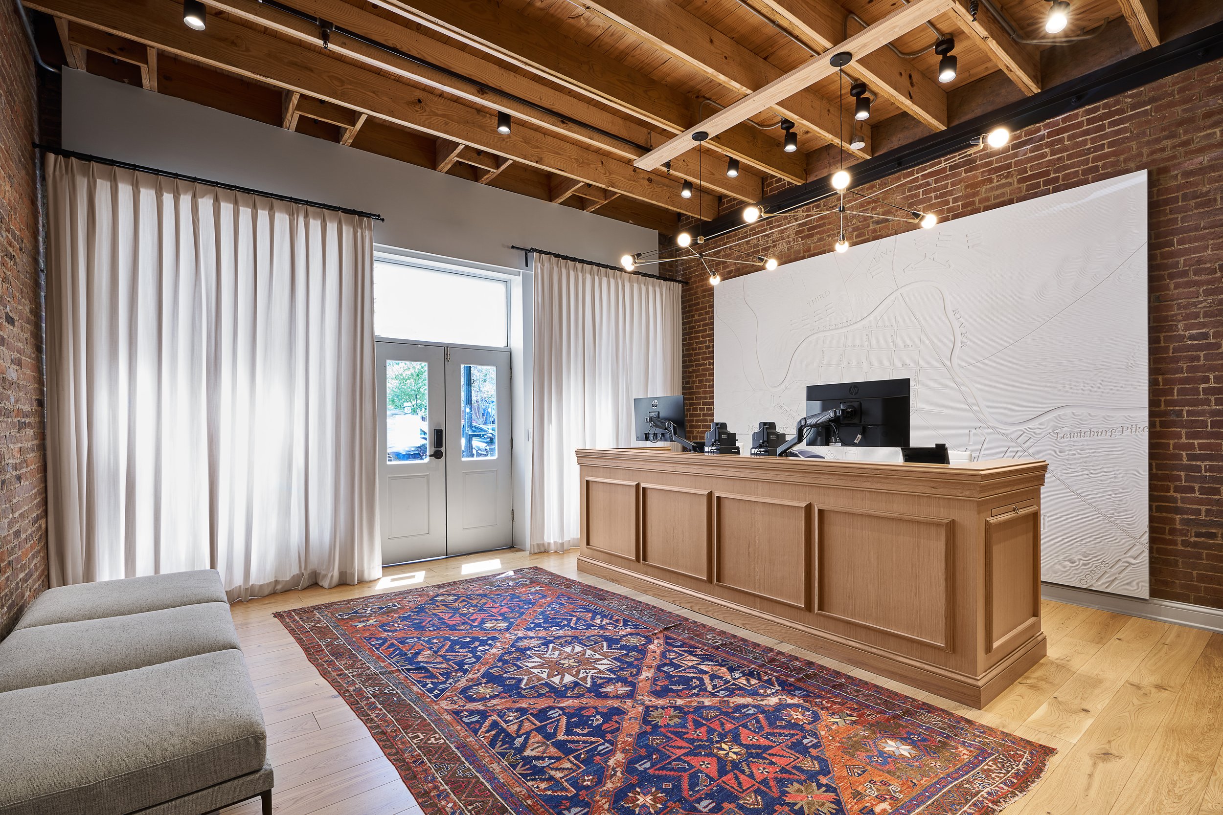Interior of a modern office with exposed brick walls, wooden ceiling beams, white curtains, a red patterned rug, a beige couch, a reception desk with computers, and a large wall map.