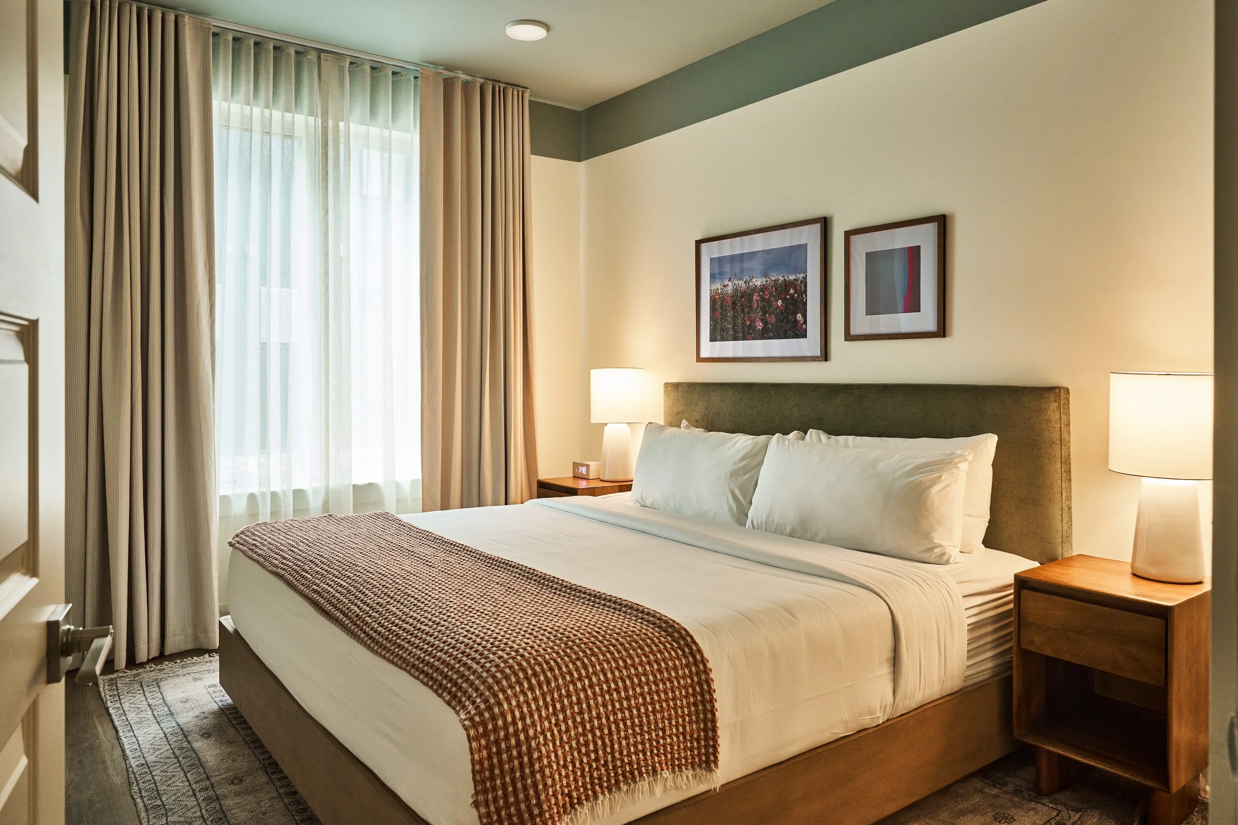 A hotel room with a neatly made bed, bedside tables with lamps, framed landscape and abstract art on the wall, and curtains over a window.