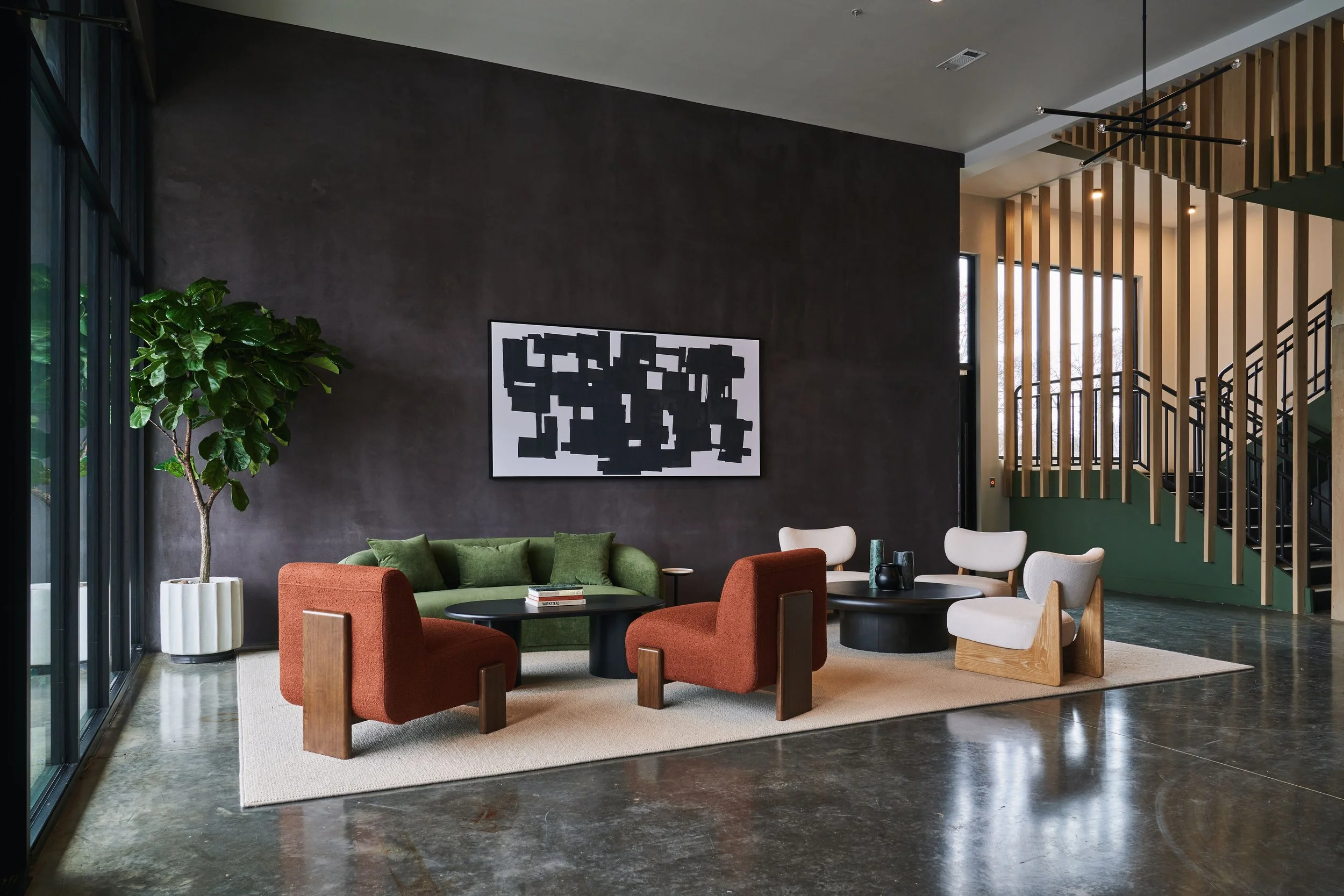 Modern living room with green, white, and rust-colored chairs around a black coffee table, a large abstract black and white artwork on a dark gray wall, a tall potted plant, large windows with black frames, wooden vertical slats near a staircase, and a beige rug on a polished concrete floor.