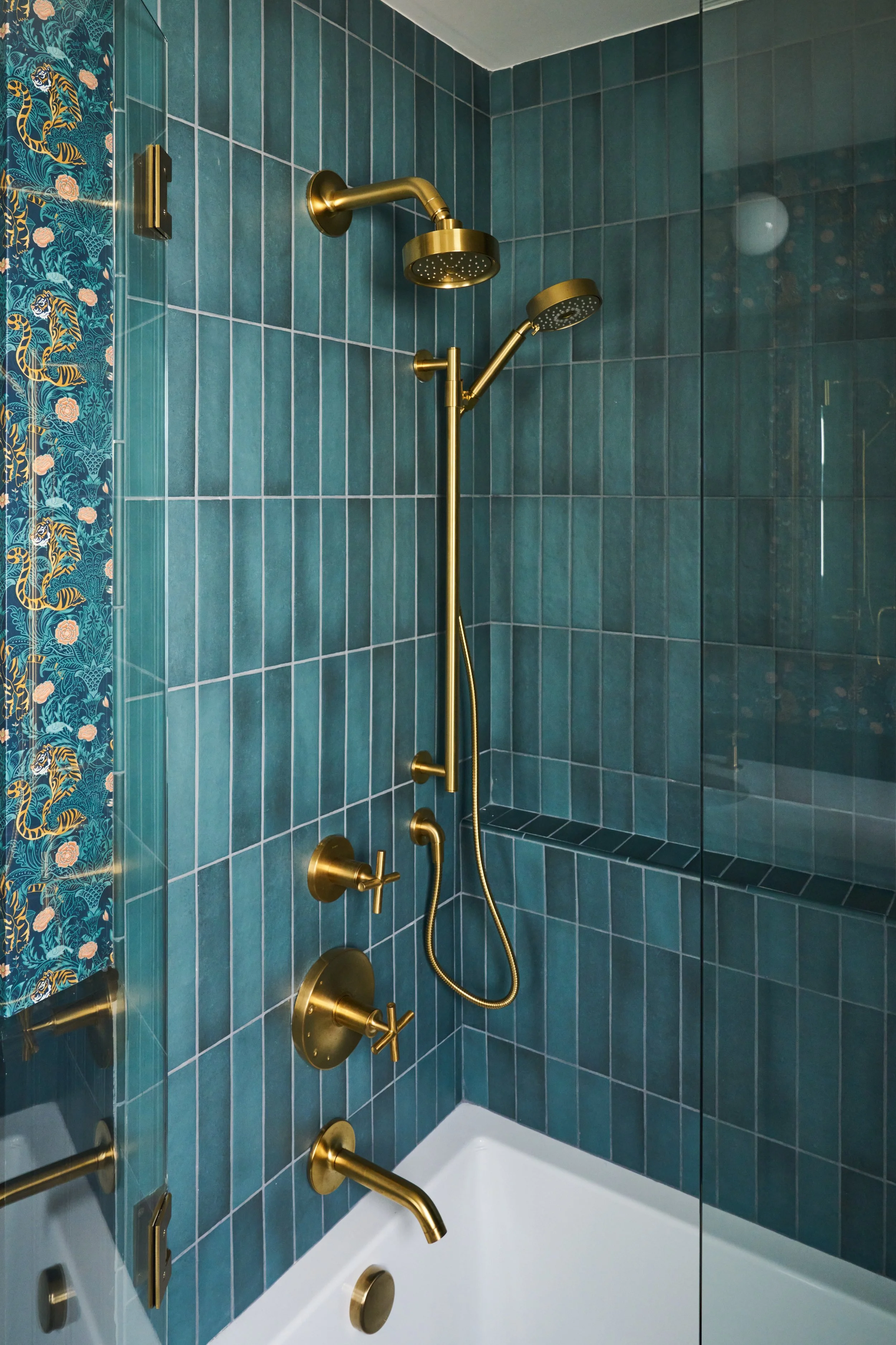 A shower with gold fixtures, including two showerheads and multiple handles, set against teal tiled walls. A glass shower door with a decorative panel featuring a tiger and floral pattern is visible on the left.
