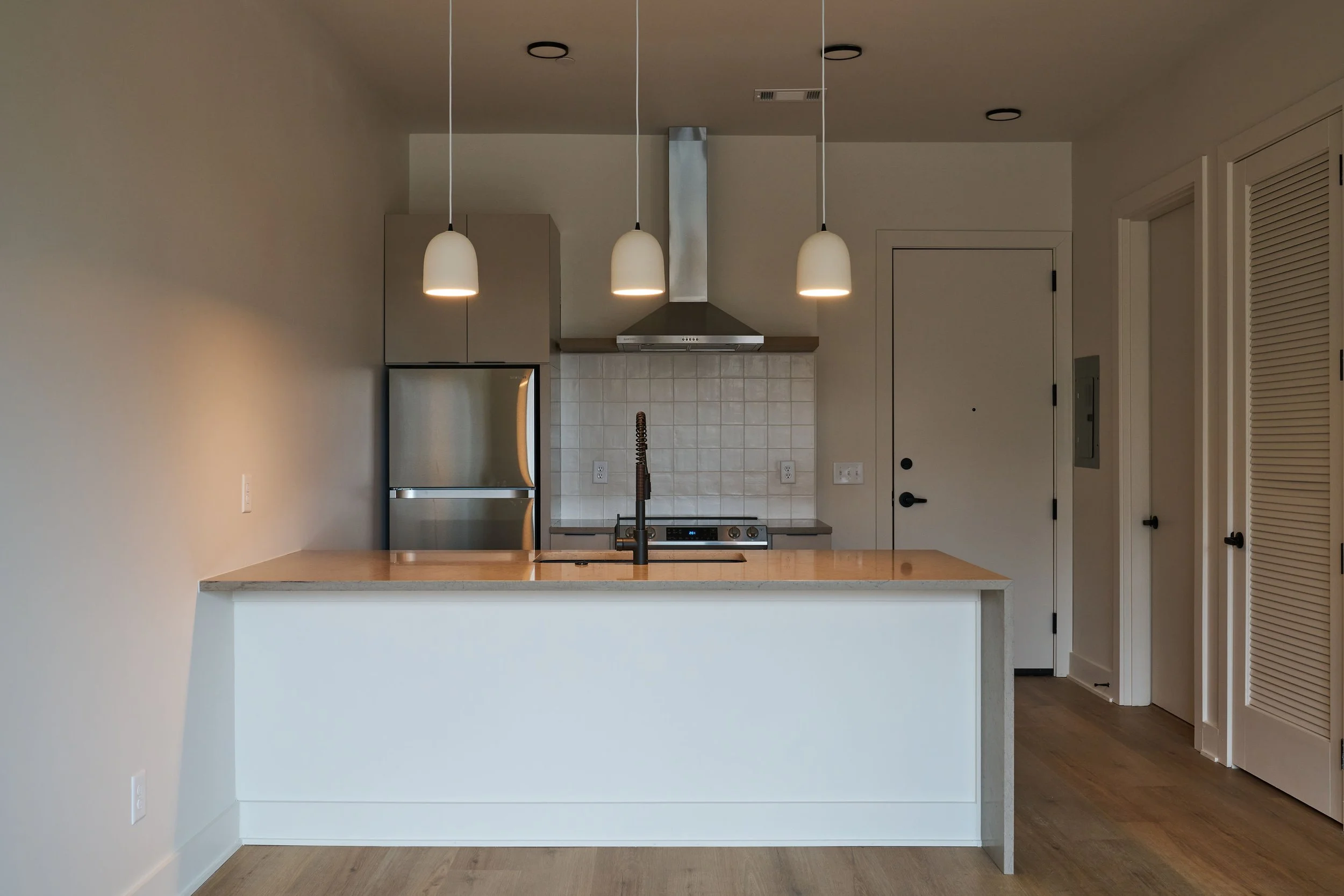 Modern kitchen with beige pendant lights over a white island, stainless steel appliances, tiled backsplash, and wood floor.