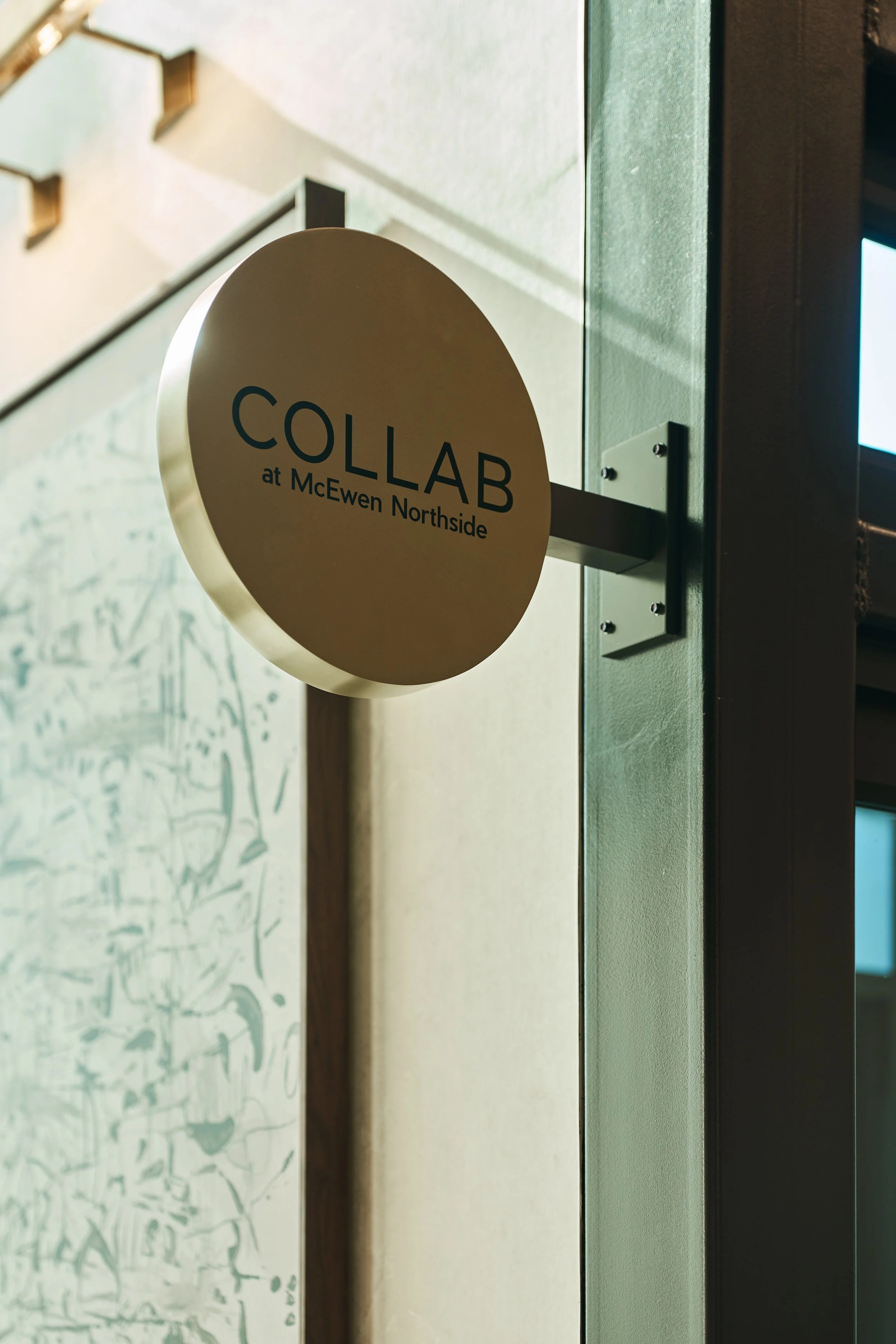 Close-up of a round sign that reads 'COLLAB at McEwen Northside,' mounted on a wall with a green color.