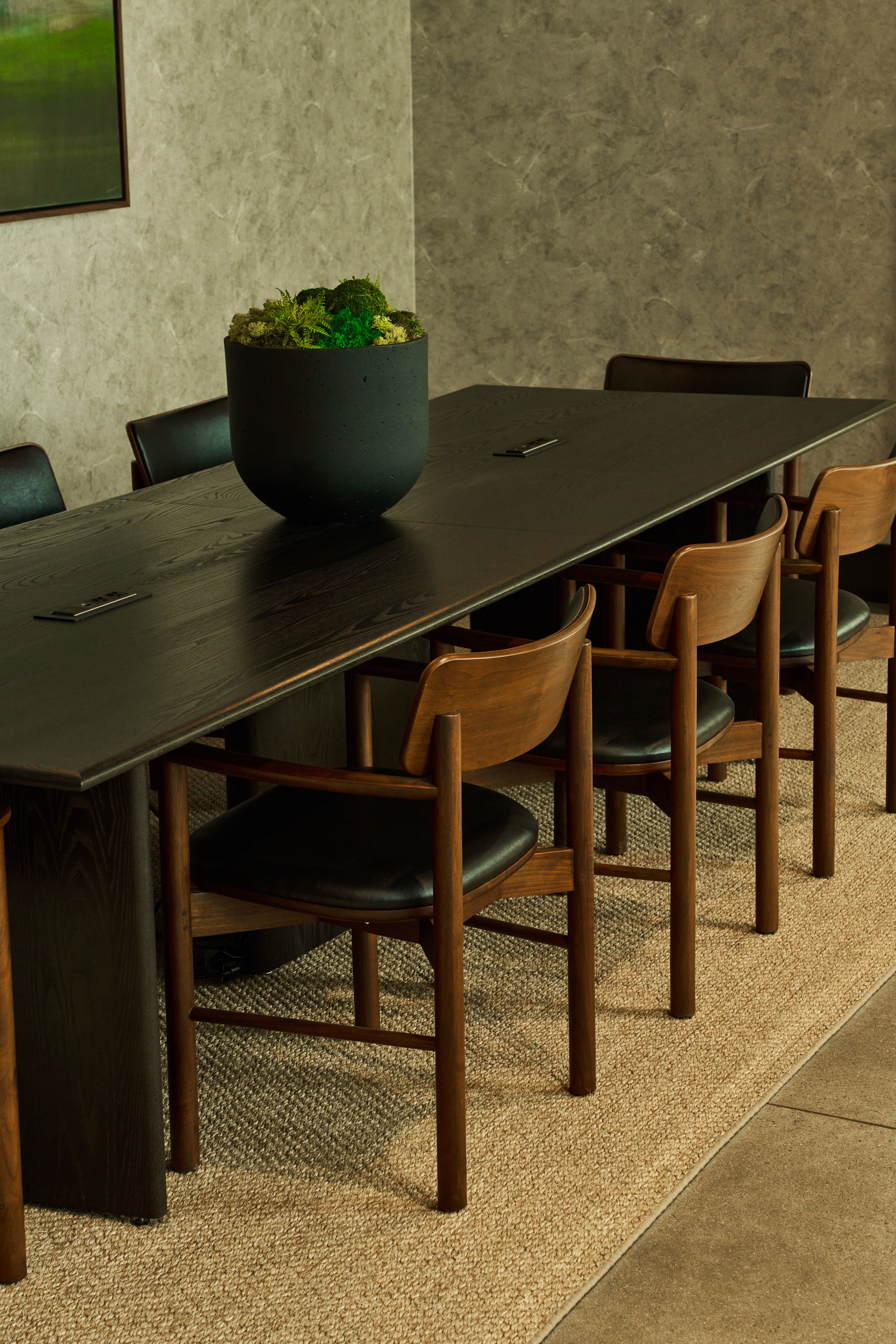 A long dark wooden table with black and wooden chairs surrounding it. A potted green plant is on the table. The room has a beige textured wall and a carpeted floor.