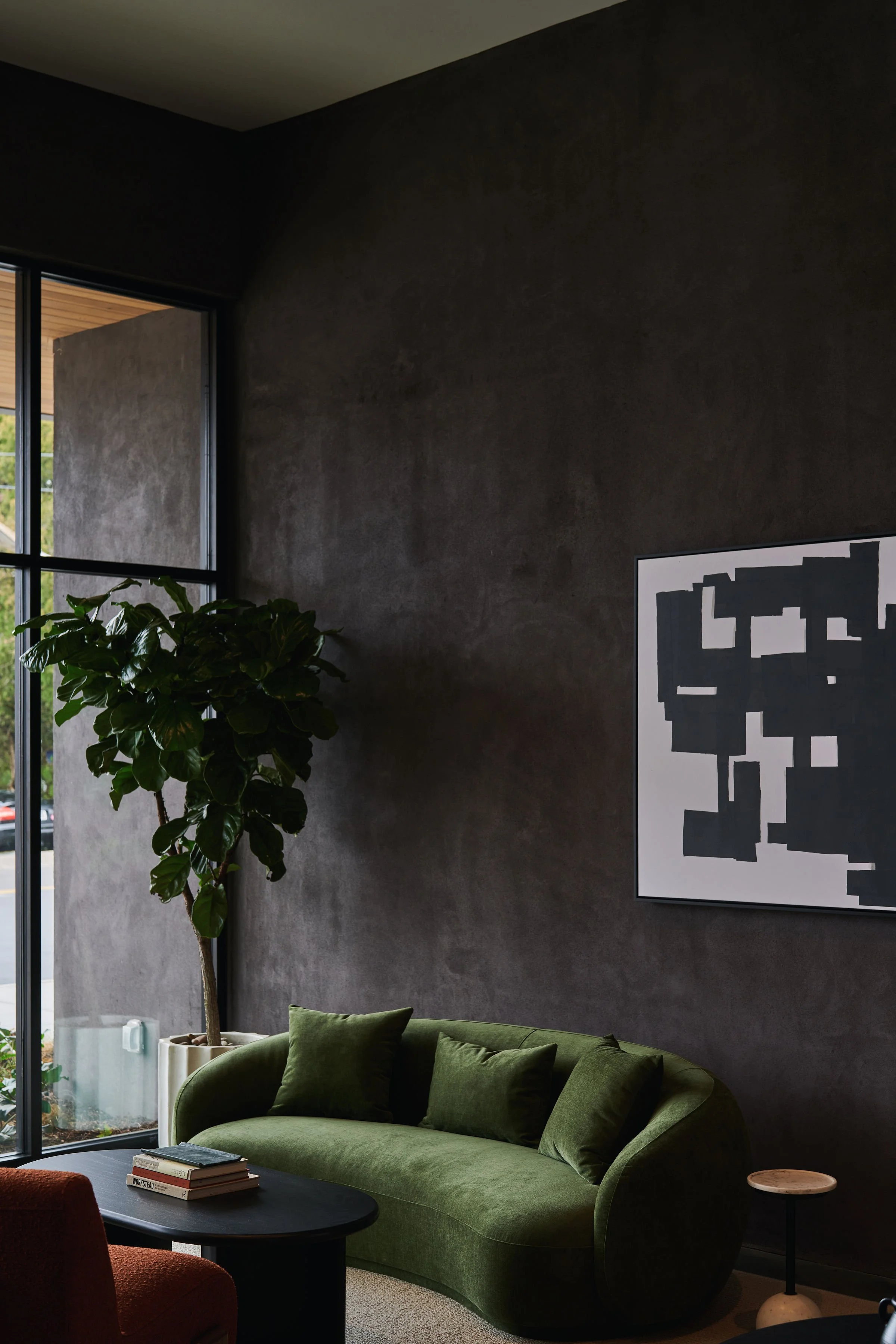 Living room corner with a dark wall, green velvet sofa, a black coffee table, a large potted plant near a floor-to-ceiling window, a black and white artwork on the wall, and a small side table with a stone top.