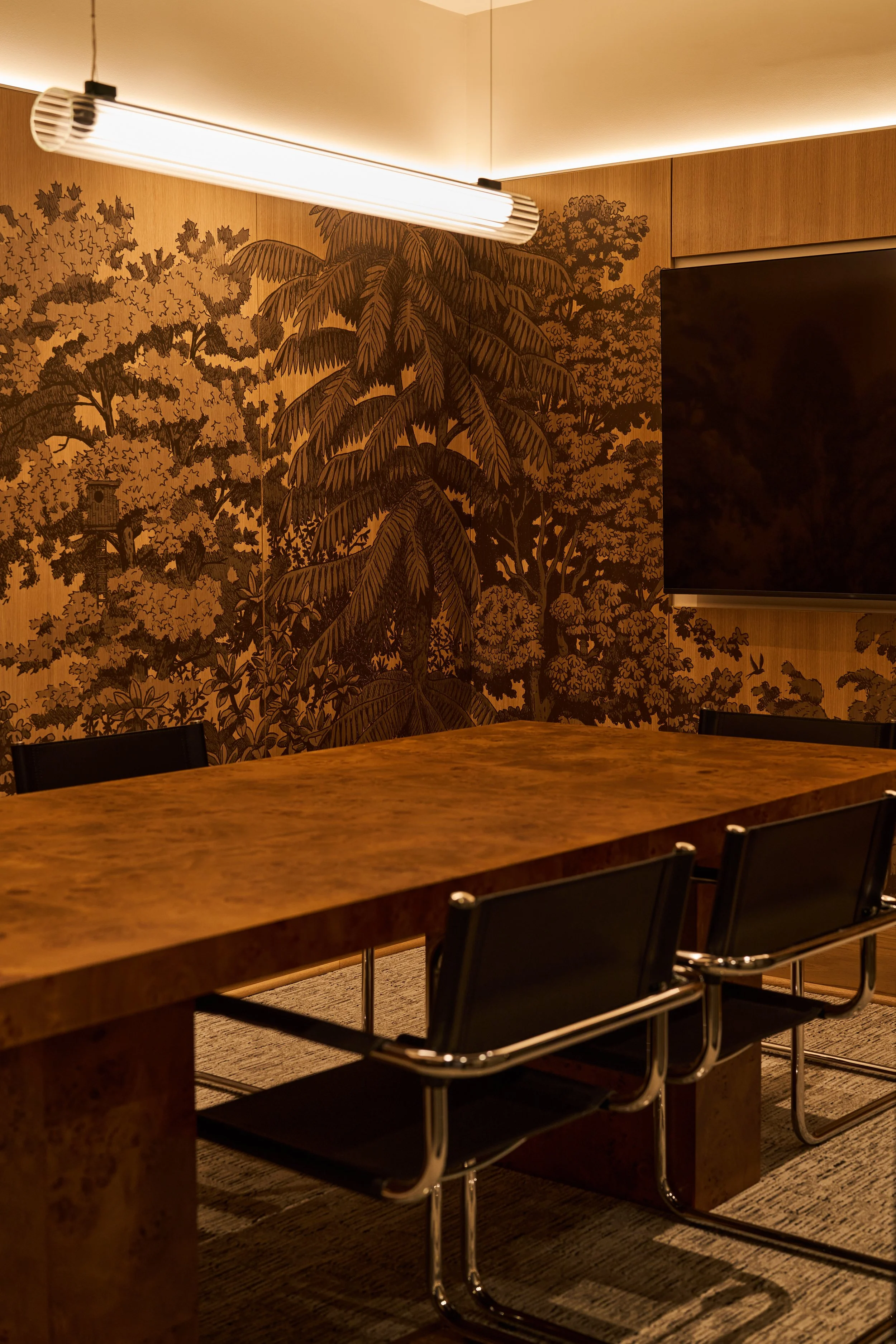 A conference room with a large wooden table, black chairs, a mural of trees on the wall, a wall-mounted flat screen TV, and a modern ceiling light fixture.