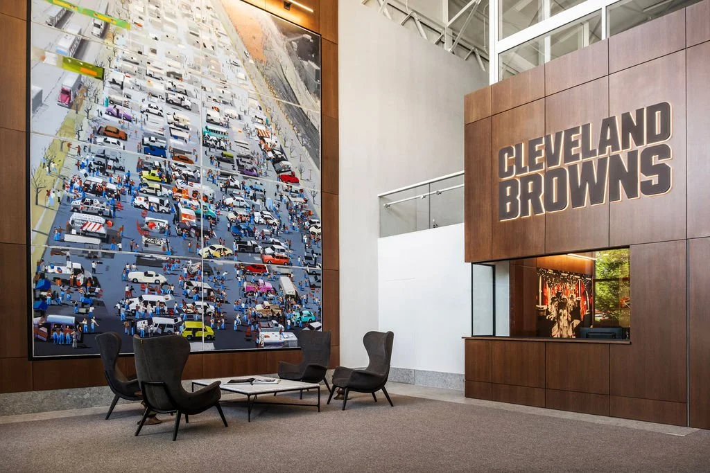 Lobby area of Cleveland Browns stadium or office with a large digital screen displaying an aerial view of a crowded parking lot and a wall with Cleveland Browns signage.