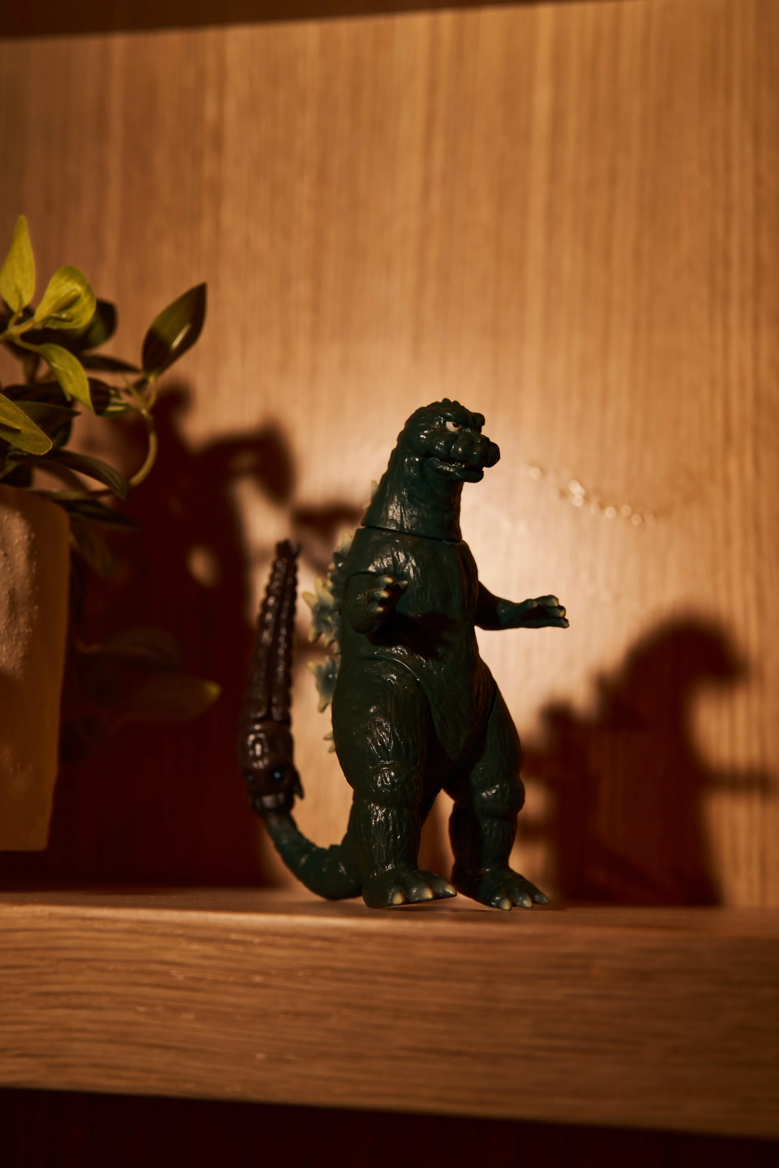 A black Godzilla toy figure standing on a wooden shelf next to a potted plant. The background features a wooden wall.