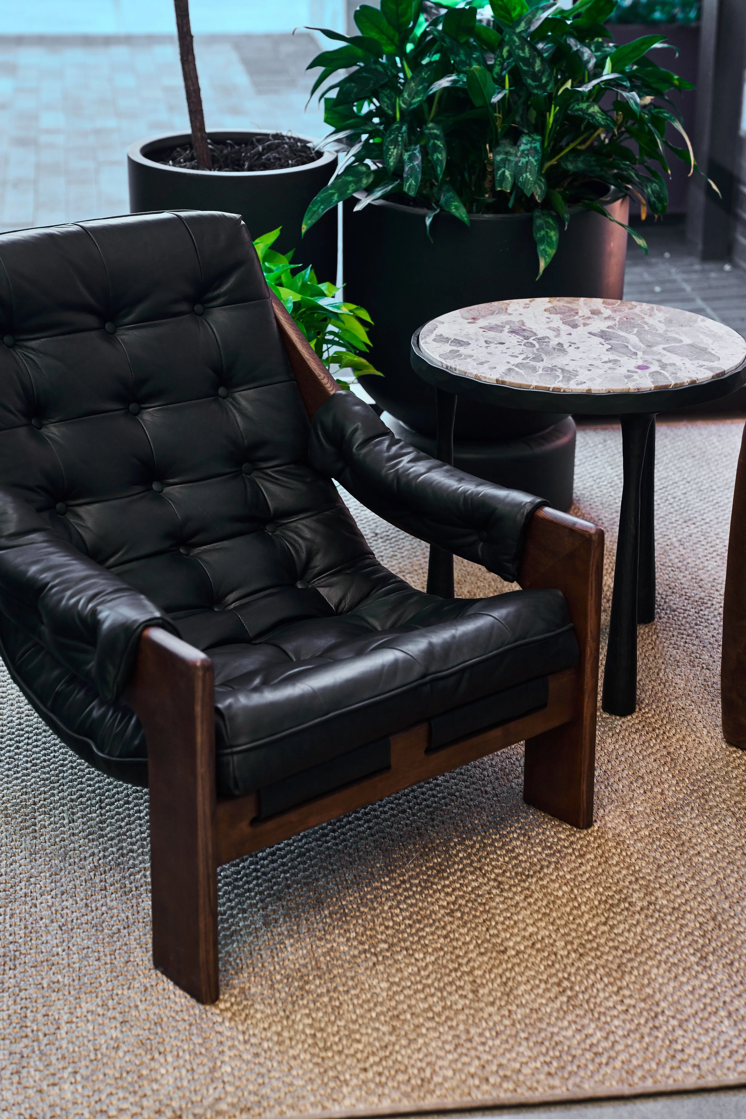 A black leather armchair with wooden frame next to a small round marble top table, with large potted plants in the background.