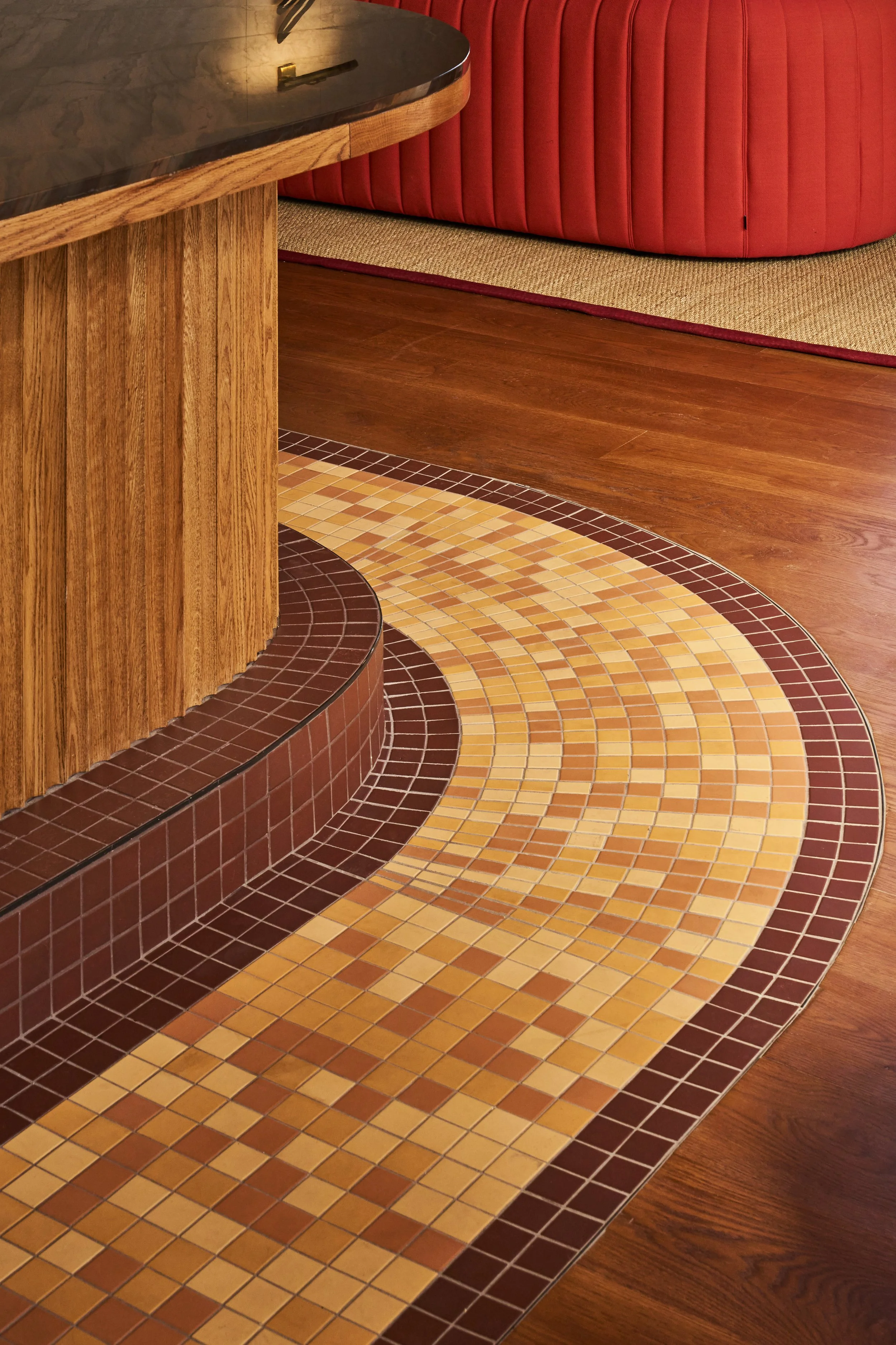 Part of a wooden counter with a dark polished surface, a red upholstered booth, and a colorful mosaic tile floor with shades of brown, yellow, and beige.