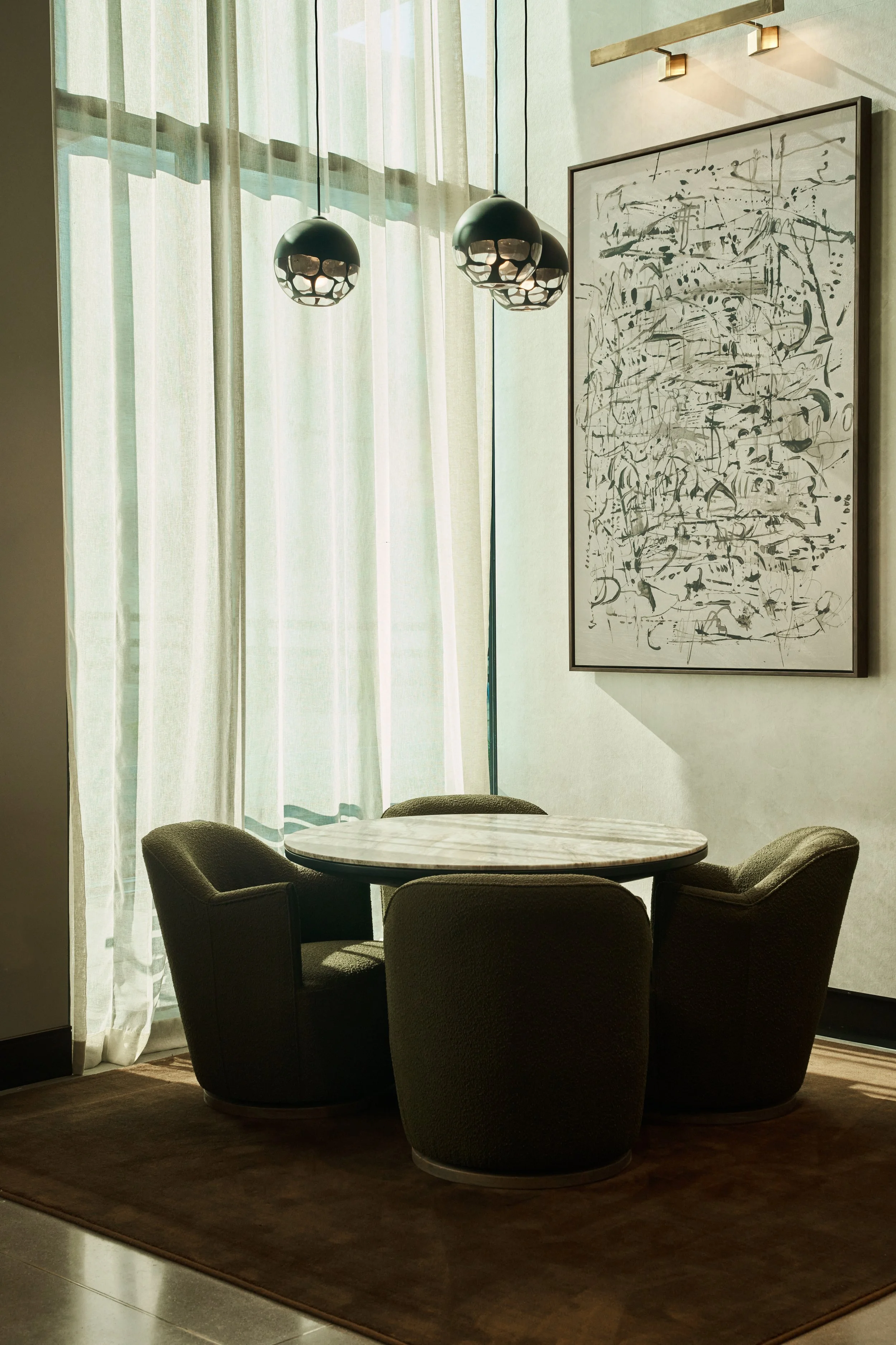 A round marble dining table surrounded by five black upholstered chairs in a modern interior with a wooden floor, large windows with white curtains, abstract black-and-white artwork, and three pendant lights hanging from the ceiling.