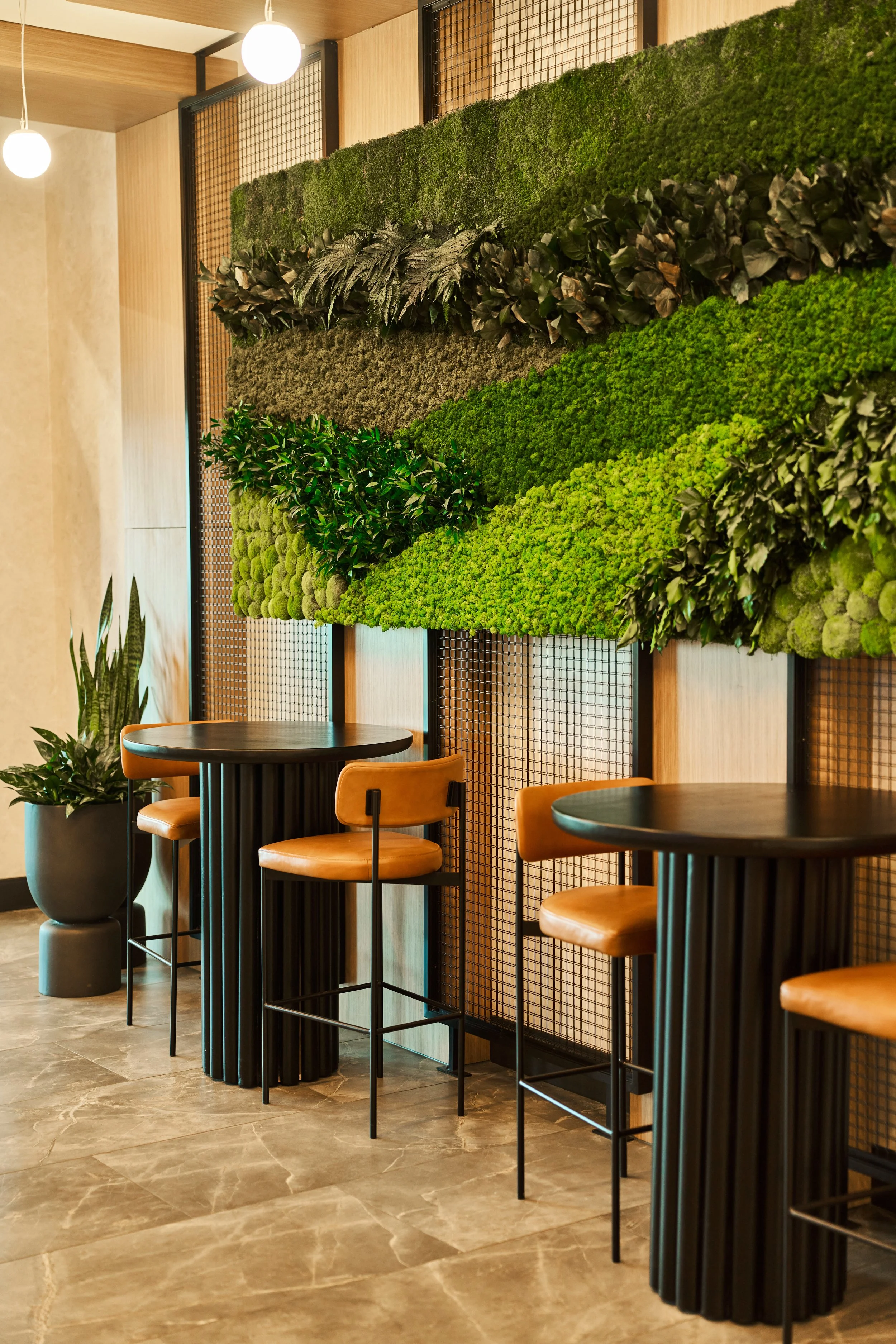 Interior of a modern café with marble tile flooring, black round tables, orange leather chairs, and a decorative green wall with various plants, illuminated by hanging spherical lights.