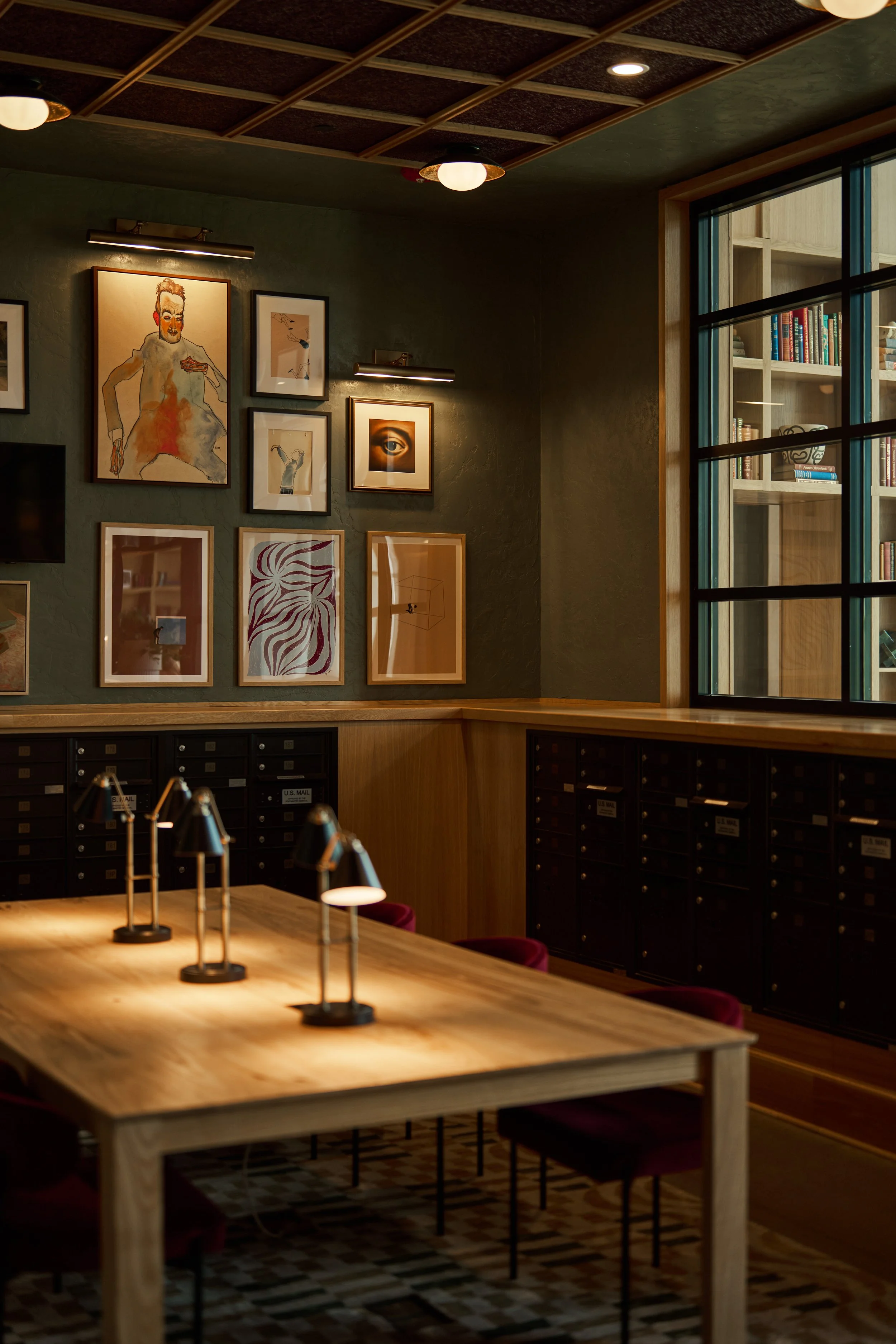 Interior of a cozy, modern library or study space with artworks on the walls, a large wooden table with lamps, and a bookshelf visible through a window.