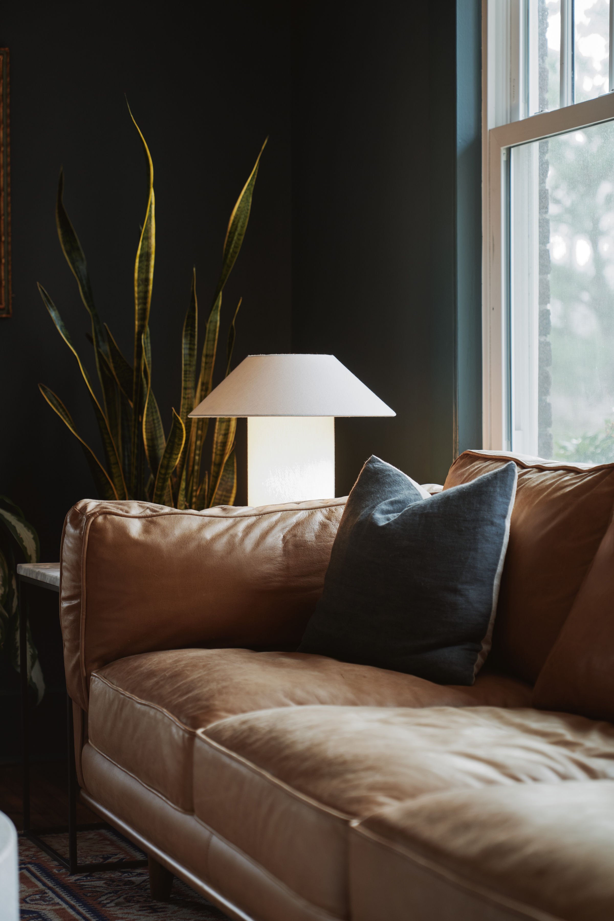 Living room with a large tan leather sofa, a dark accent wall, a tall green houseplant, a white table lamp, and a window allowing natural light.