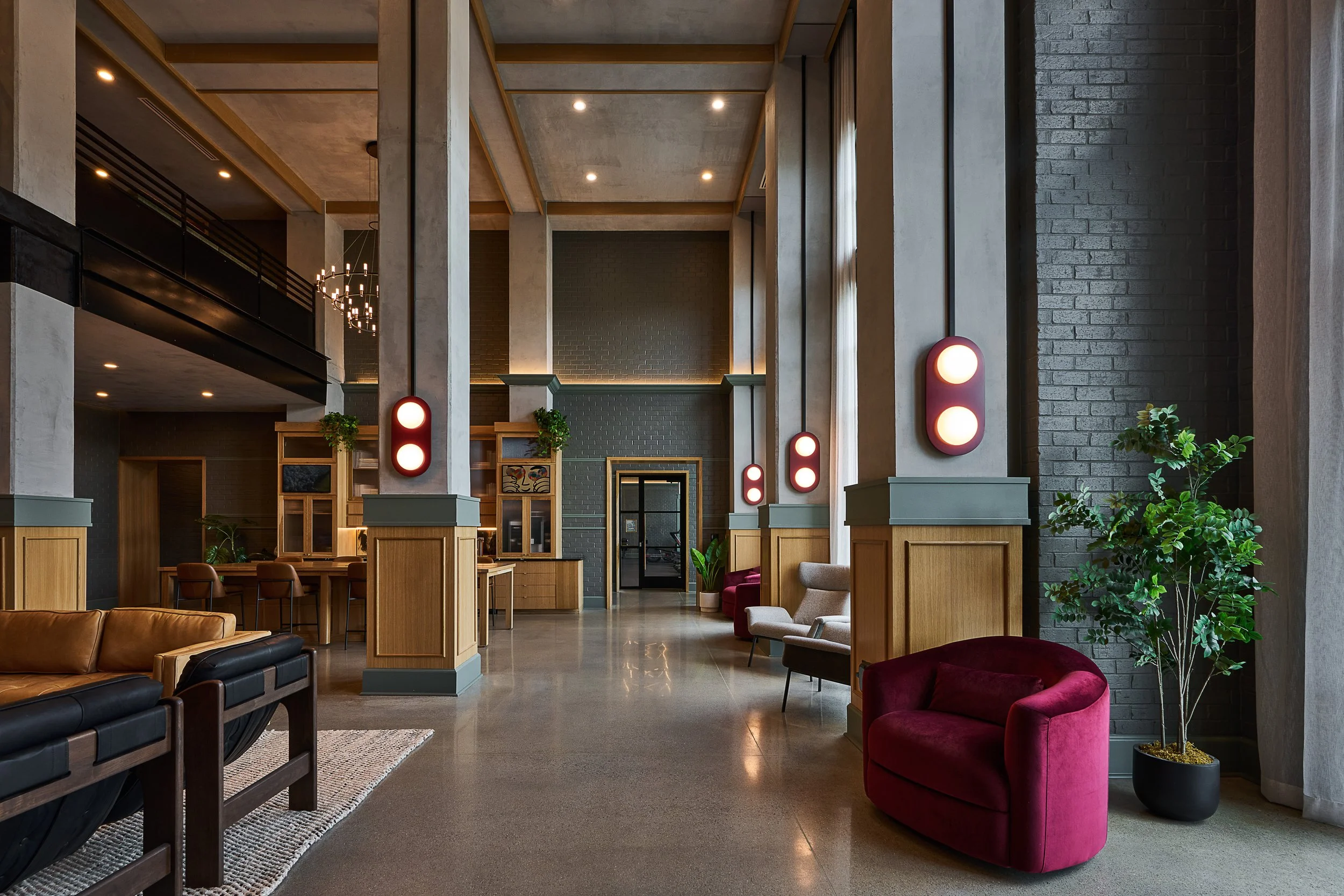 Modern high-ceiling lobby with concrete and brick walls, wood accents, and contemporary lighting. Seating includes red armchairs and sofas, with green plants for decor.