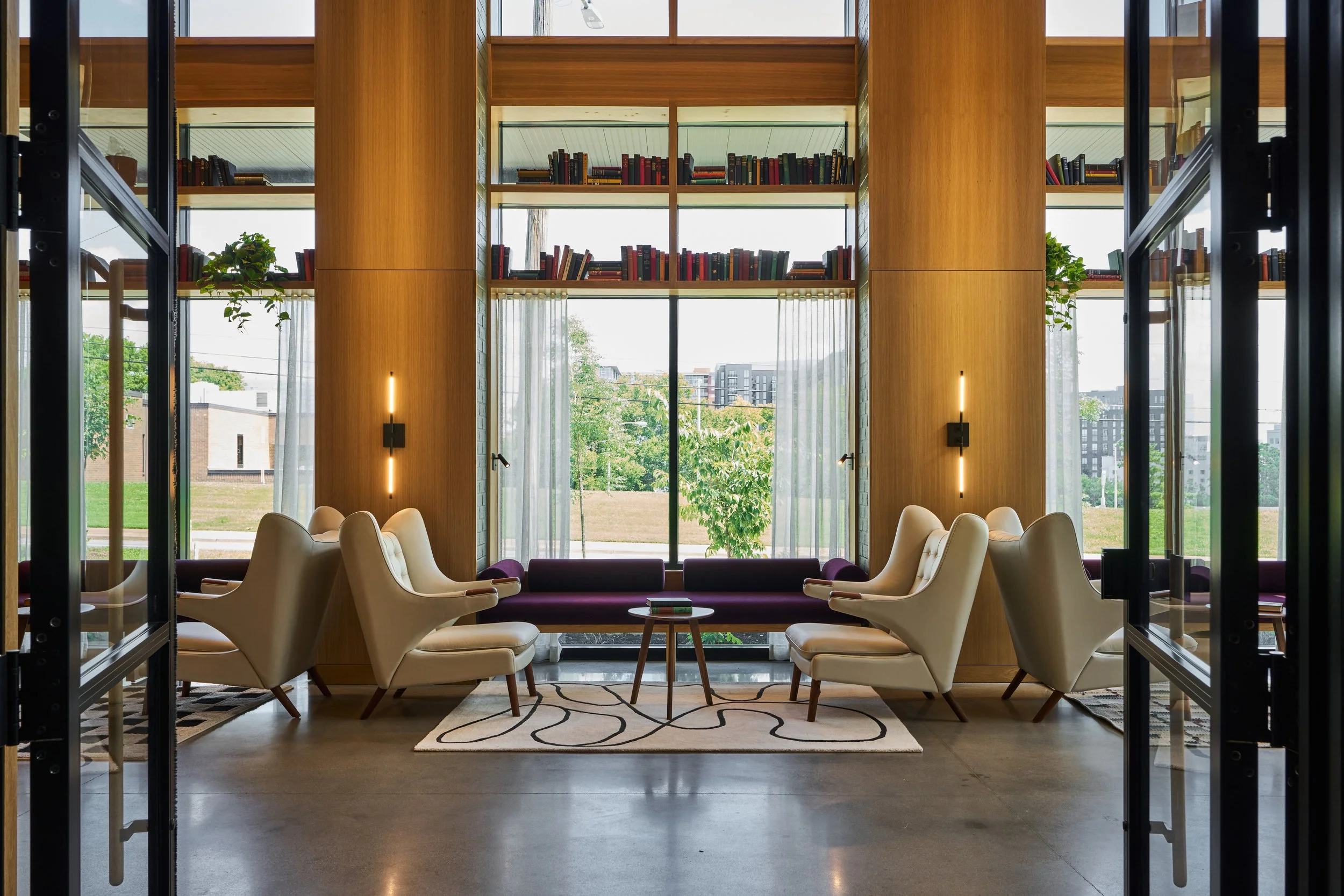 Modern lounge area with white armchairs and purple benches, large windows with sheer curtains, wooden walls, and bookshelves with books, with an outdoor view of trees and buildings.