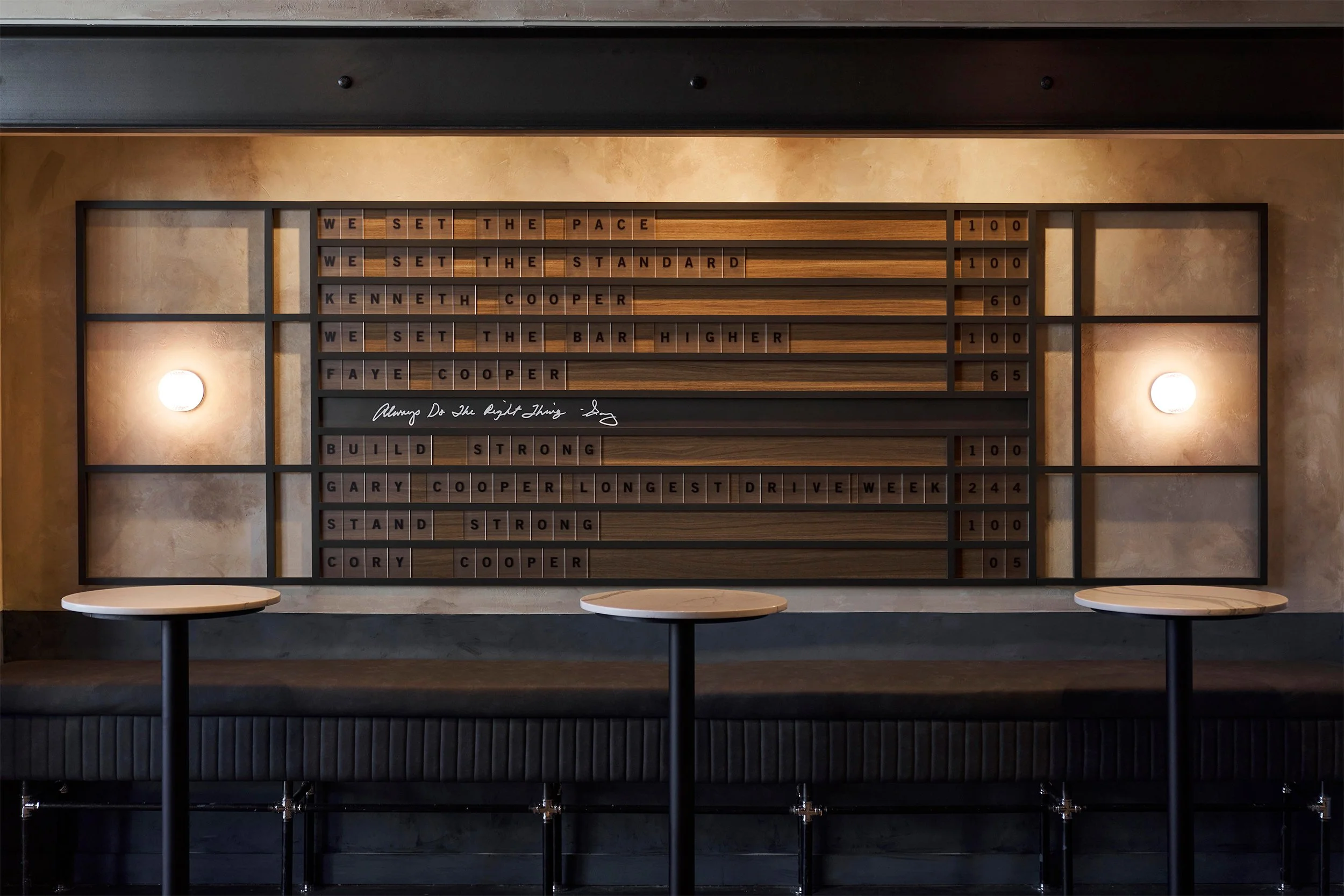 A black framed wall with wooden board game tiles displaying a motivational message, lit by two wall sconces, with two round tables in front.