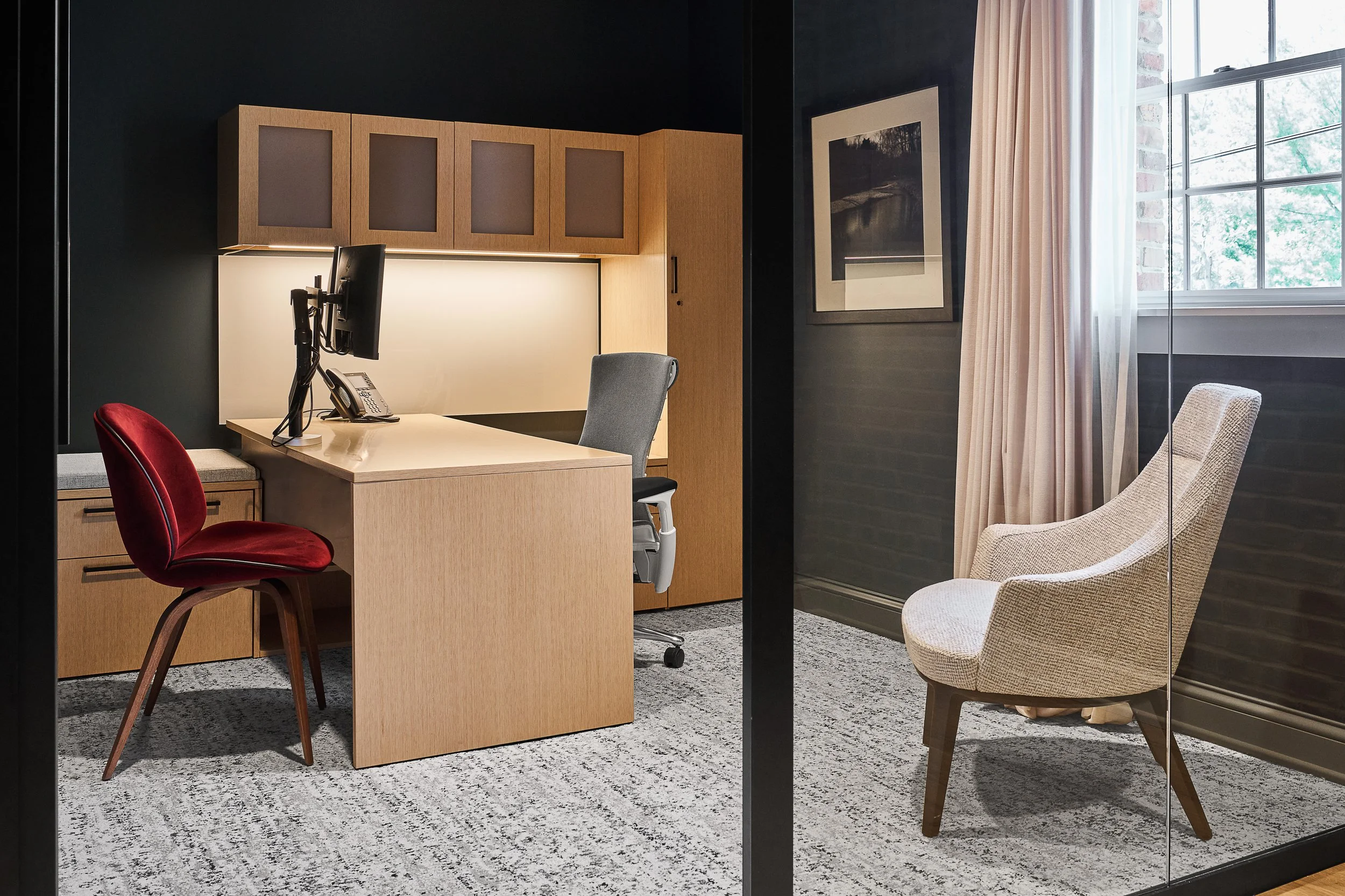 Modern office with a wooden desk, black computer monitor, telephone, red and gray chairs, a framed black-and-white photo on a dark wall, a large window with beige curtains, and a patterned gray carpet.