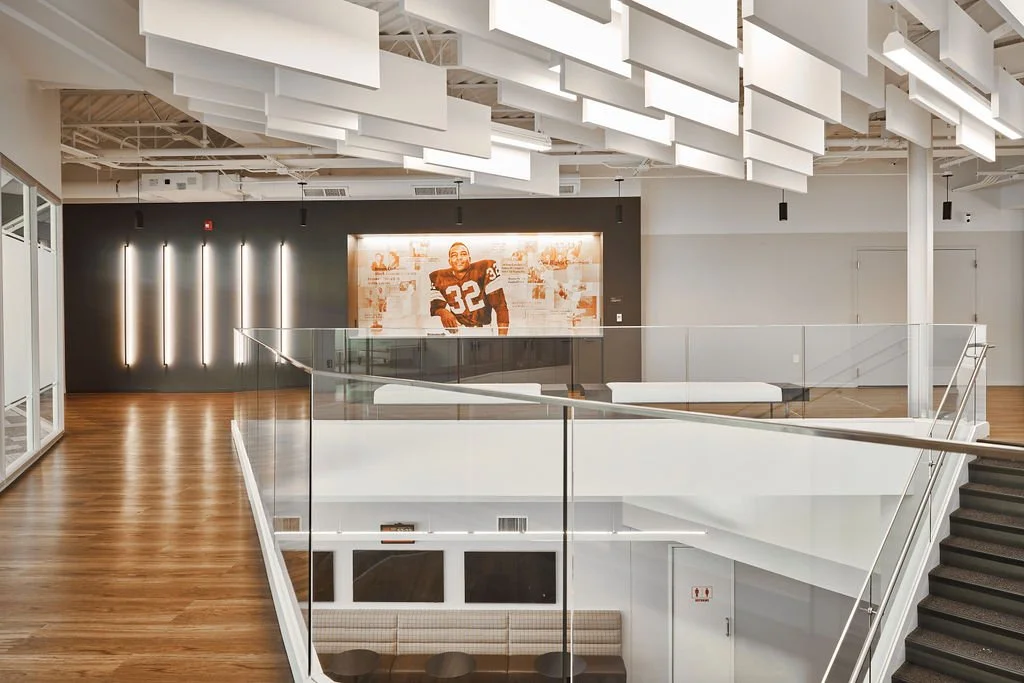 An indoor space with modern decor, featuring wood flooring, a glass railing, and abstract white ceiling panels. There is a large framed photograph of a football player wearing jersey number 32 on a dark wall.
