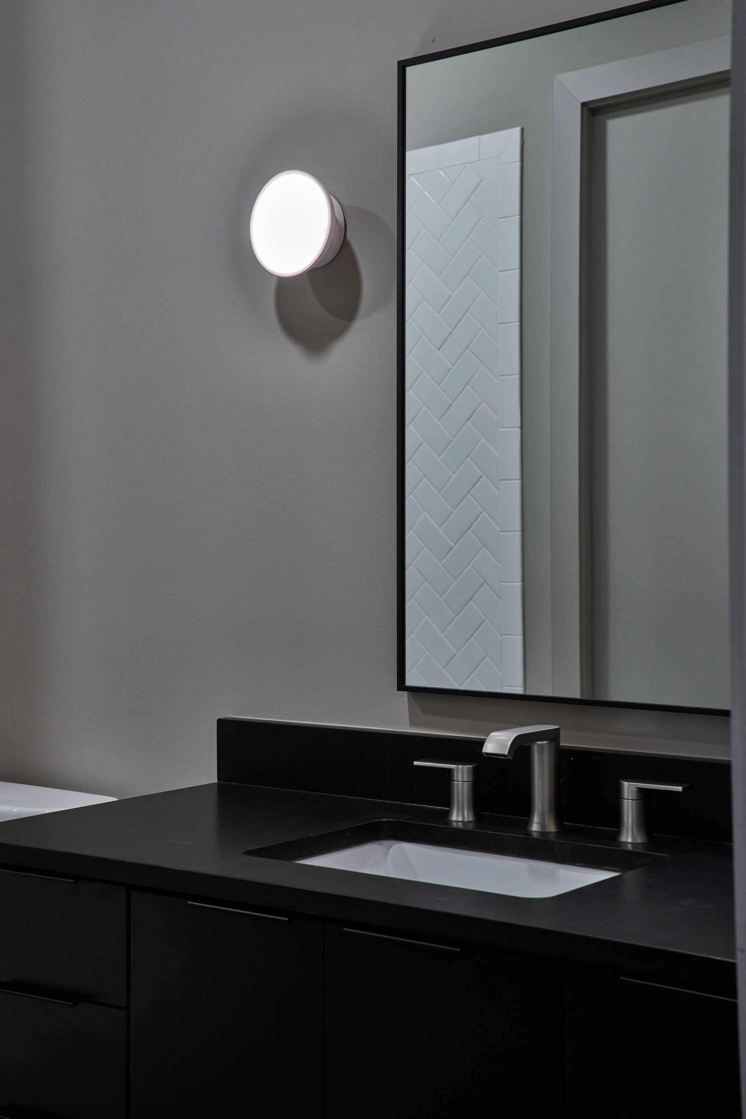 Bathroom vanity with black countertop, silver faucet, and a large rectangular mirror. Wall light fixture above the mirror and white tiled wall with a herringbone pattern reflected in the mirror.