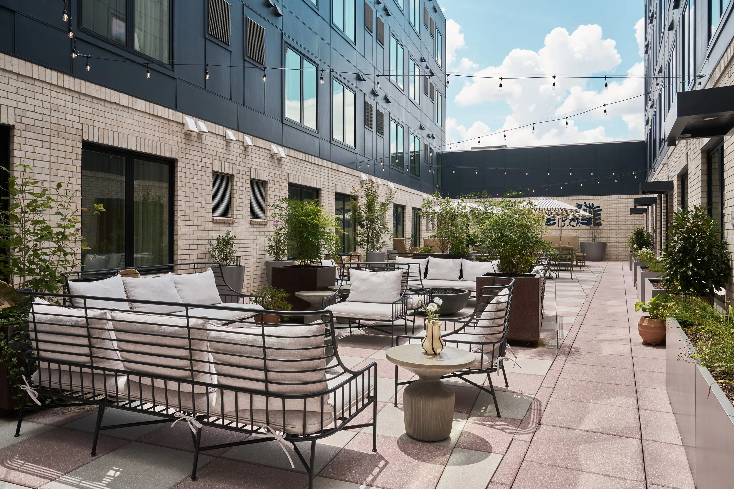 An outdoor patio area with modern black metal furniture with white cushions and a variety of potted plants. String lights hang overhead, and the background features a tall modern building with large windows, brick and dark panel exterior, under a par