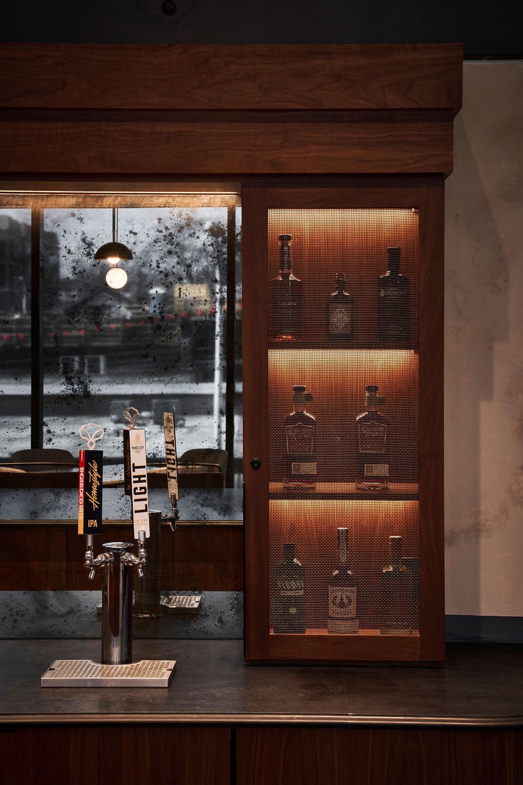 A bar with taps for craft beers in front, a window with raindrops, a mirror, and wooden shelves with bottles backlit with warm light.