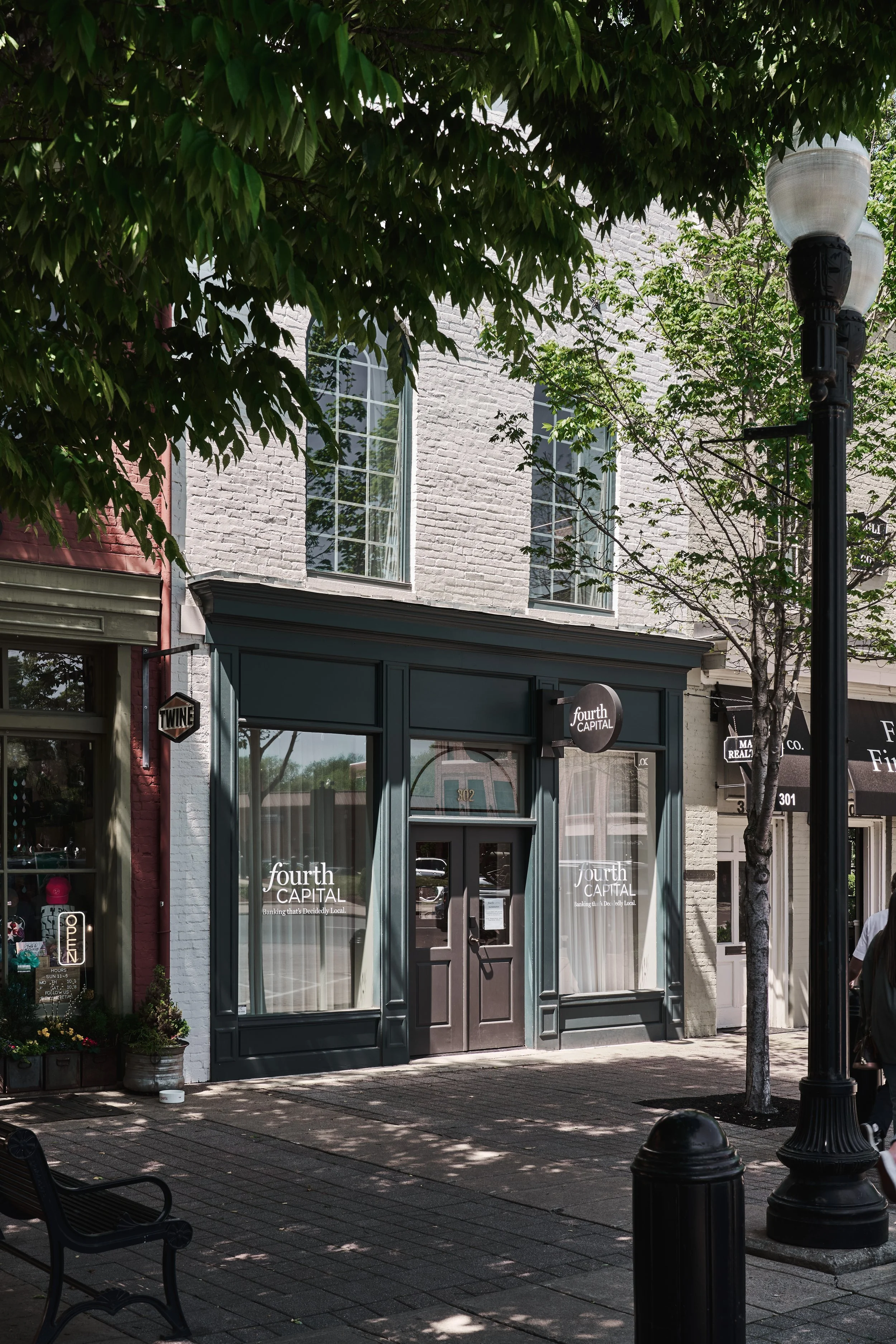 Street view showing a storefront with signage reading 'fourth CAPITAL' and large windows, surrounded by trees and street lamps, with a bench on the sidewalk.
