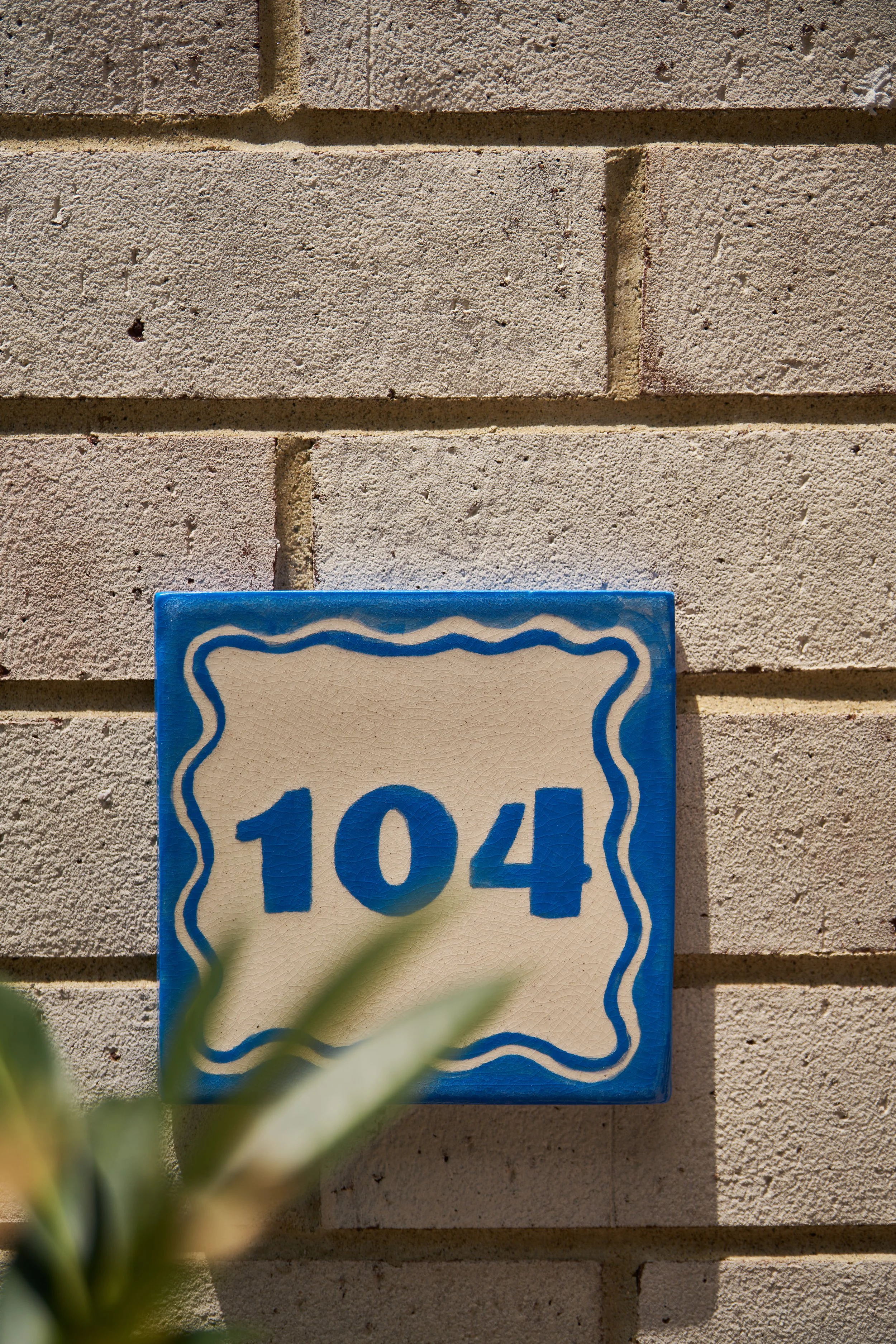 Close-up of a rectangular blue and white house number plaque with the number 104, mounted on a beige brick wall, with some greenery in the foreground.