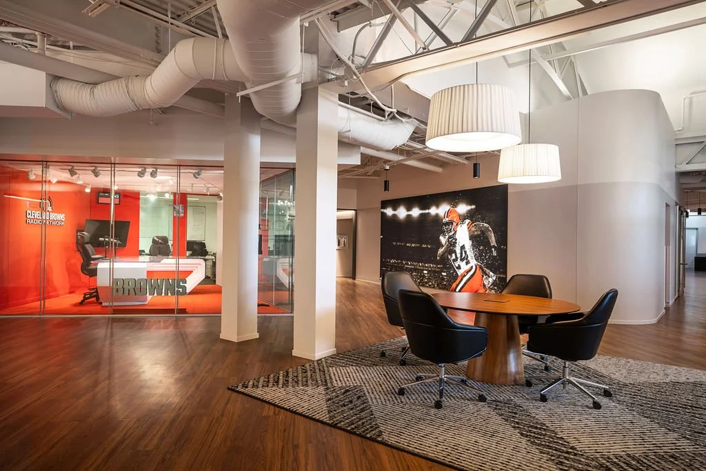 Office space with a round wooden table and four black chairs, a large Cleveland Browns logo painting, and a glass-walled room with a reception desk and office chairs inside. Modern lighting and wooden flooring.