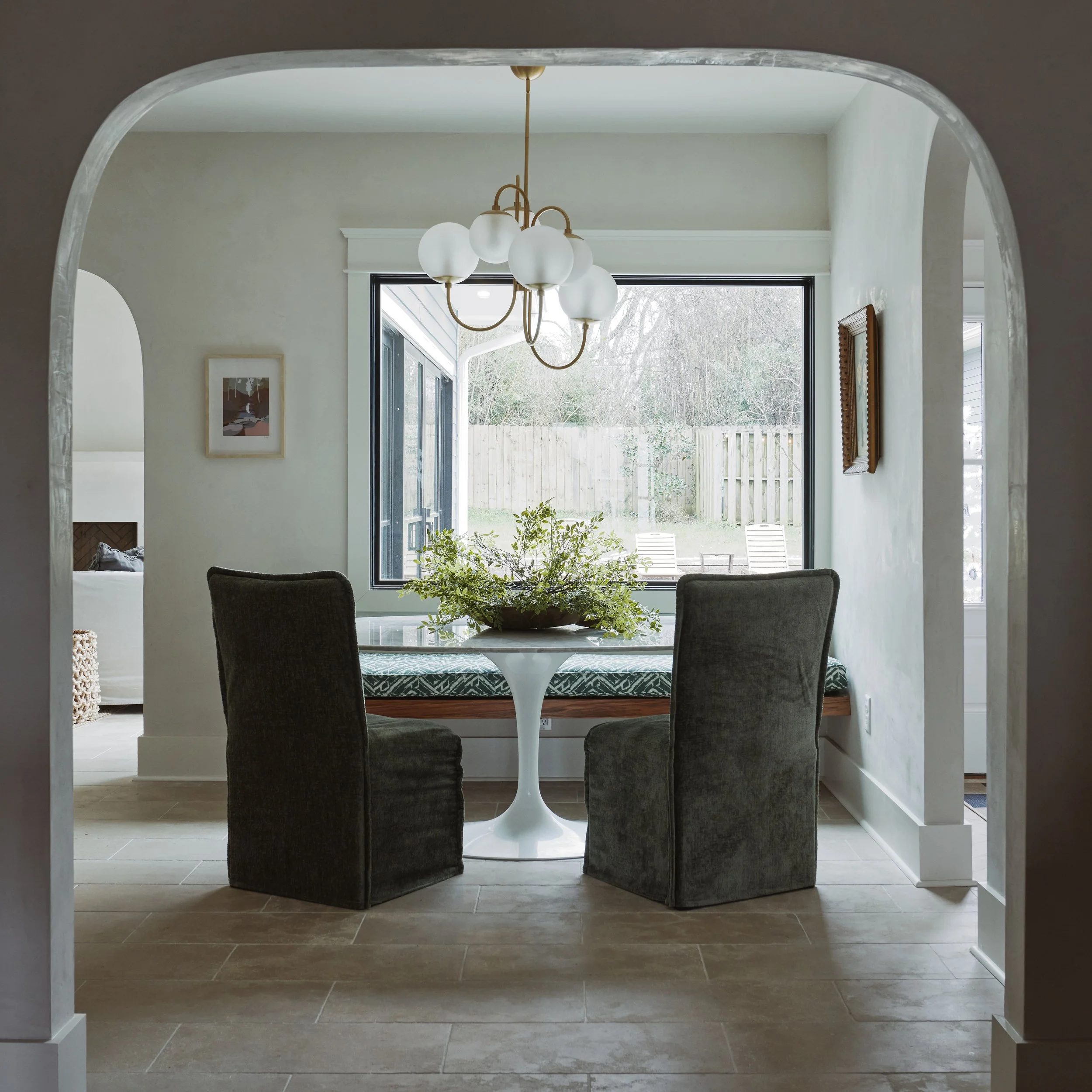 Interior view of a dining area with a round table, four dark upholstered chairs, a bowl with greenery, and a modern chandelier hanging from the ceiling. Large window shows a backyard with trees and a wooden fence.