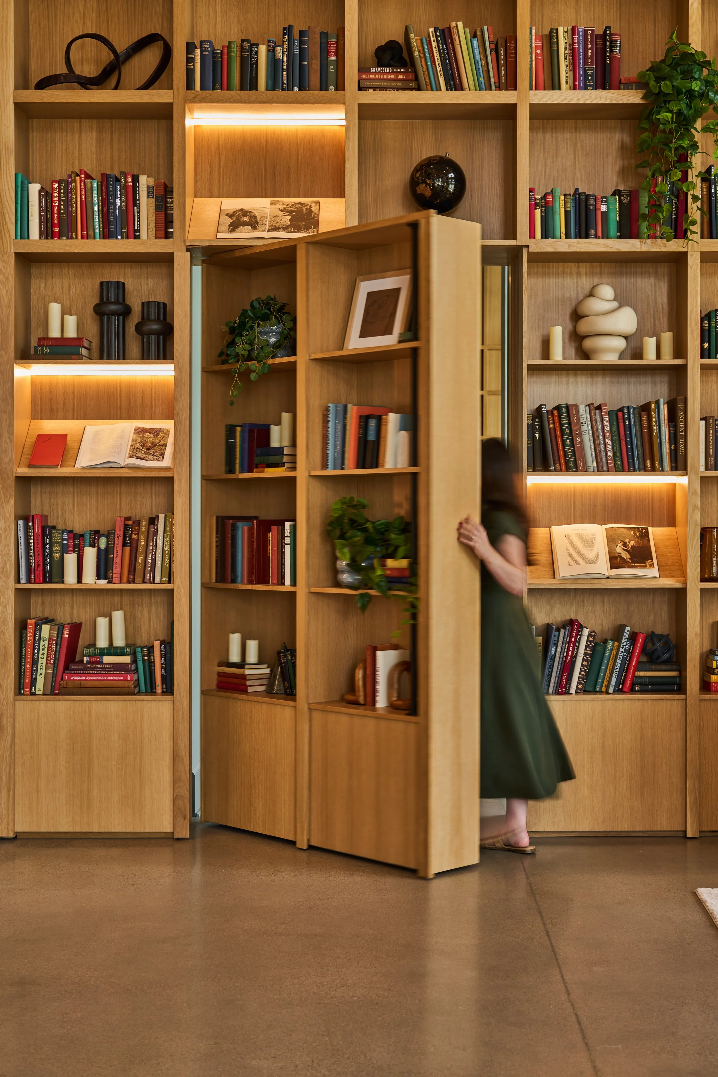 A person peeking out from behind a wooden library bookshelf, which contains various books, decorations, and plants, in a cozy bookstore or library setting.