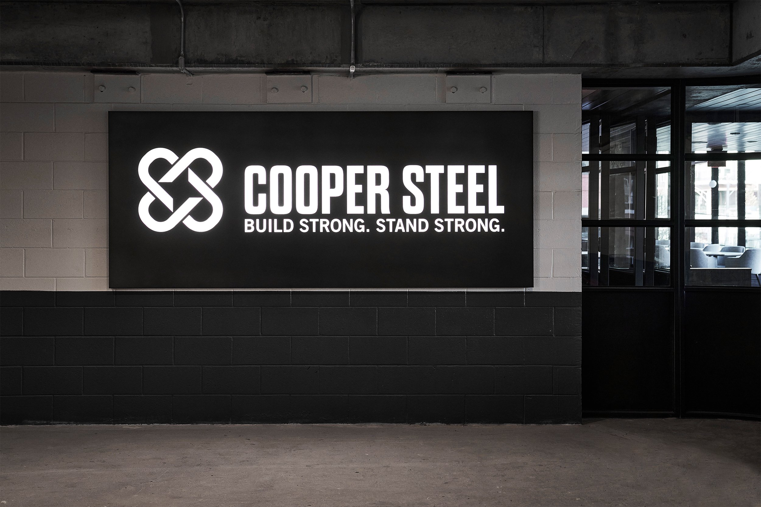 Illuminated sign reading 'COUPER STEEL BUILD STRONG. STAND STRONG.' on a black wall in an industrial interior with large windows.