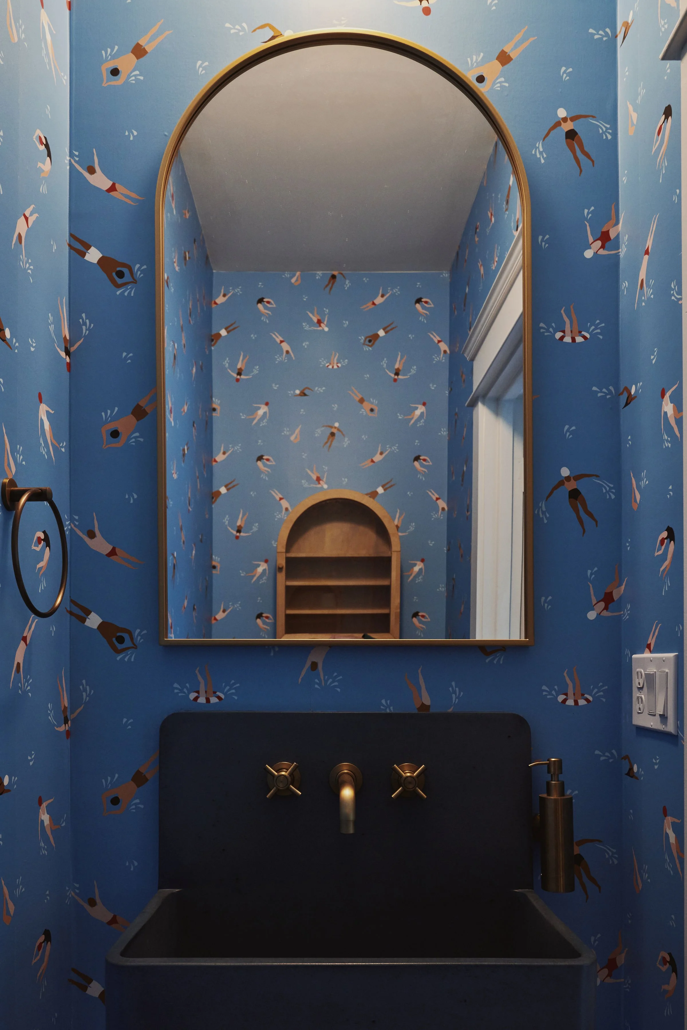 Bathroom vanity with a large mirror, light blue wallpaper with illustrated swimmers and water-related figures, black countertop with gold fixtures, a soap dispenser, and a towel ring.