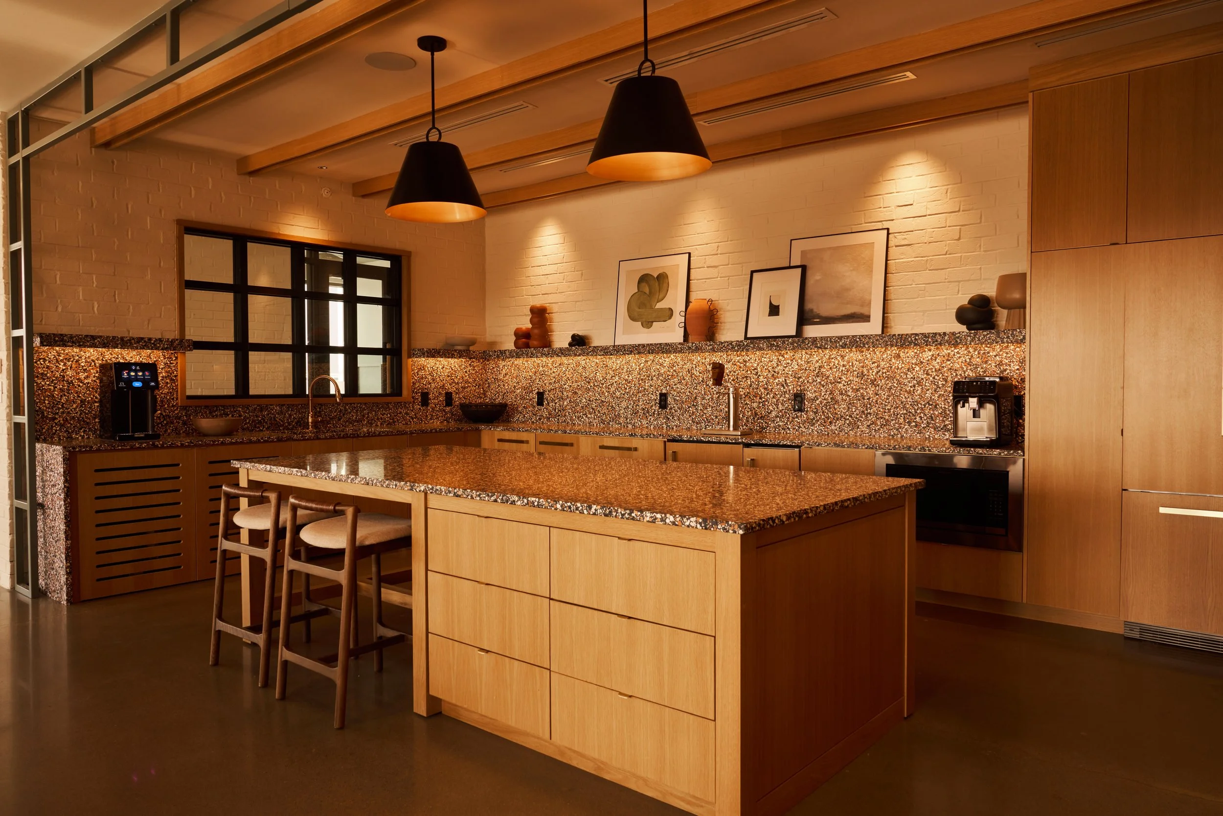 A modern kitchen with light wood cabinets, a granite countertop, and black light fixtures. Decor includes framed abstract and landscape art, ceramic vases, and a coffee machine. There are chairs around a kitchen island and a large window.