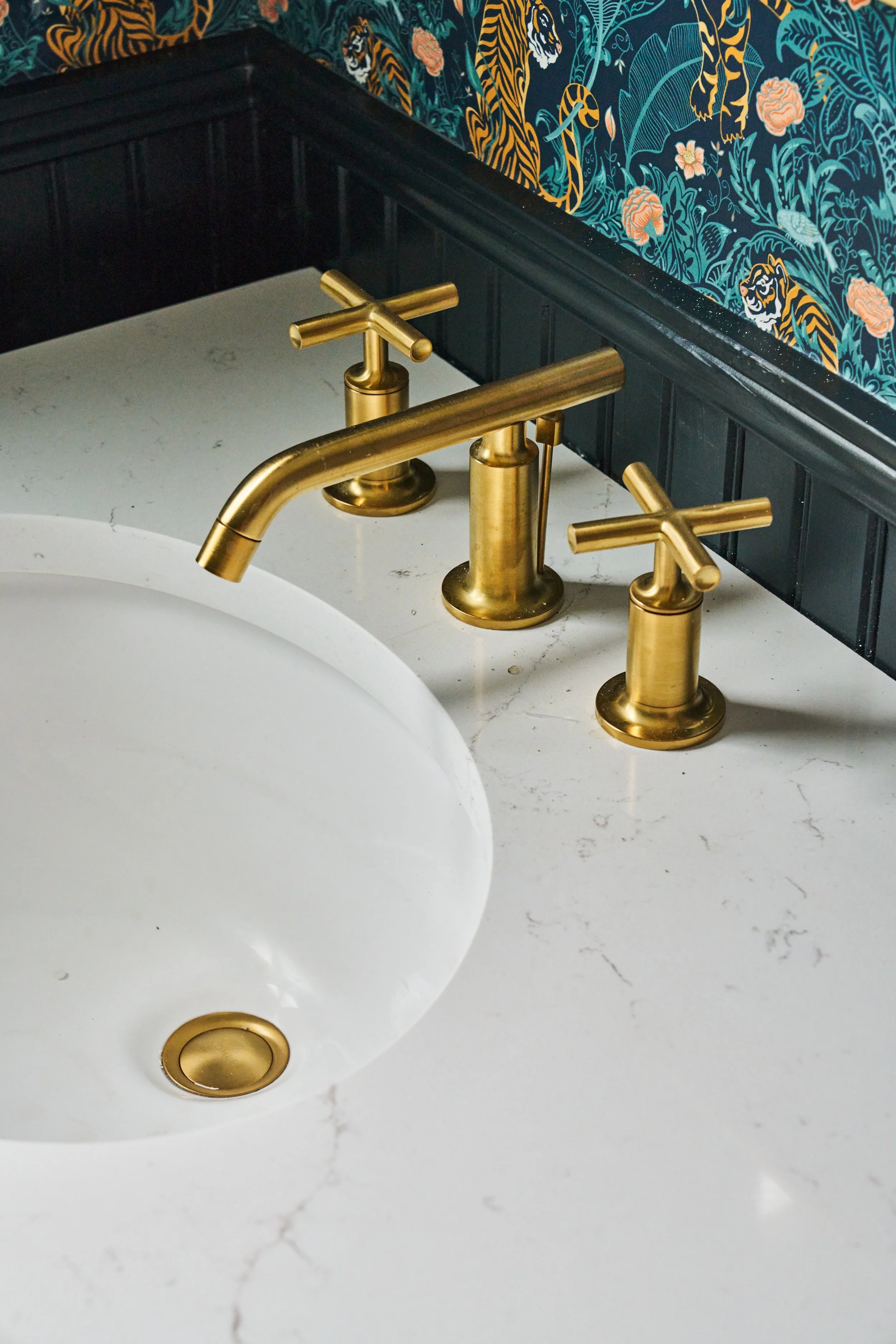 Close-up of a white bathroom sink with a gold faucet and handles, set against a black wainscoting wall and patterned wallpaper featuring tigers, flowers, and foliage.