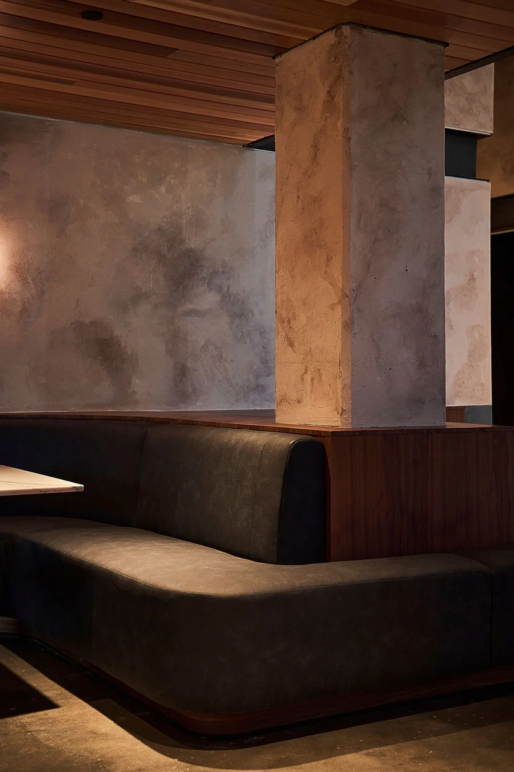 Interior of a modern restaurant with a curved dark booth, concrete walls, wooden ceiling, and a partial wooden divider.
