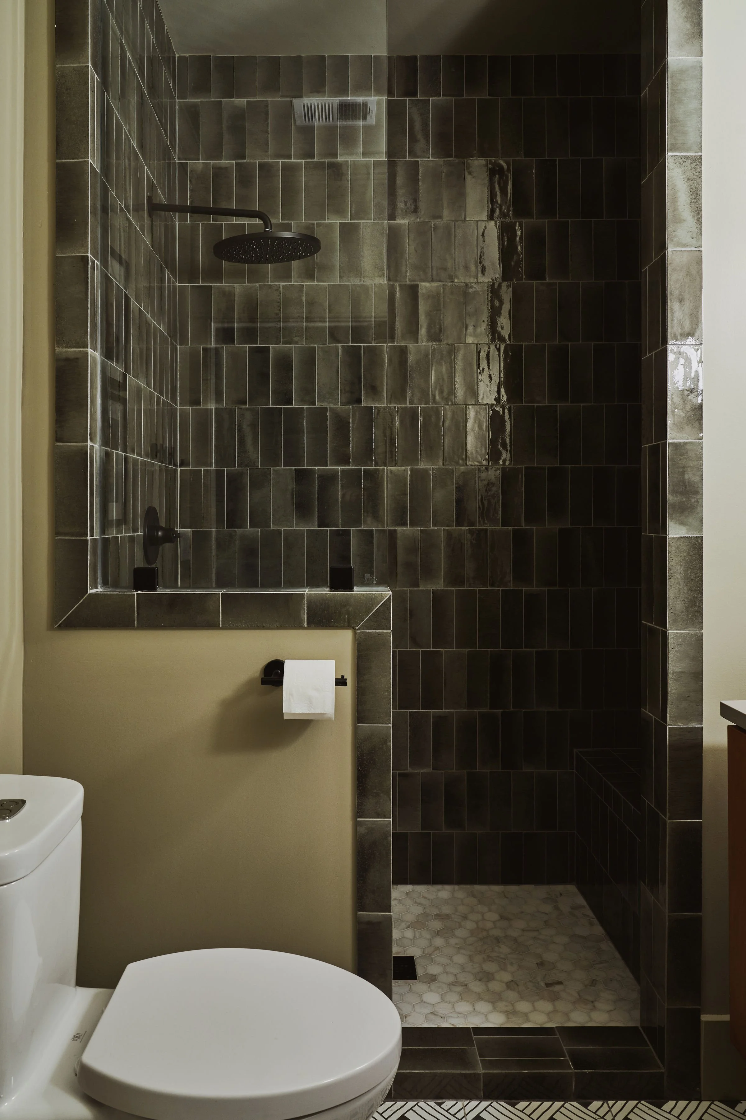 A modern bathroom with a white toilet in the foreground, beige wall, a towel holder with a toilet paper roll, a dark tiled walk-in shower with a black showerhead, black fixtures, and a mix of hexagon and rectangular floor tiles.