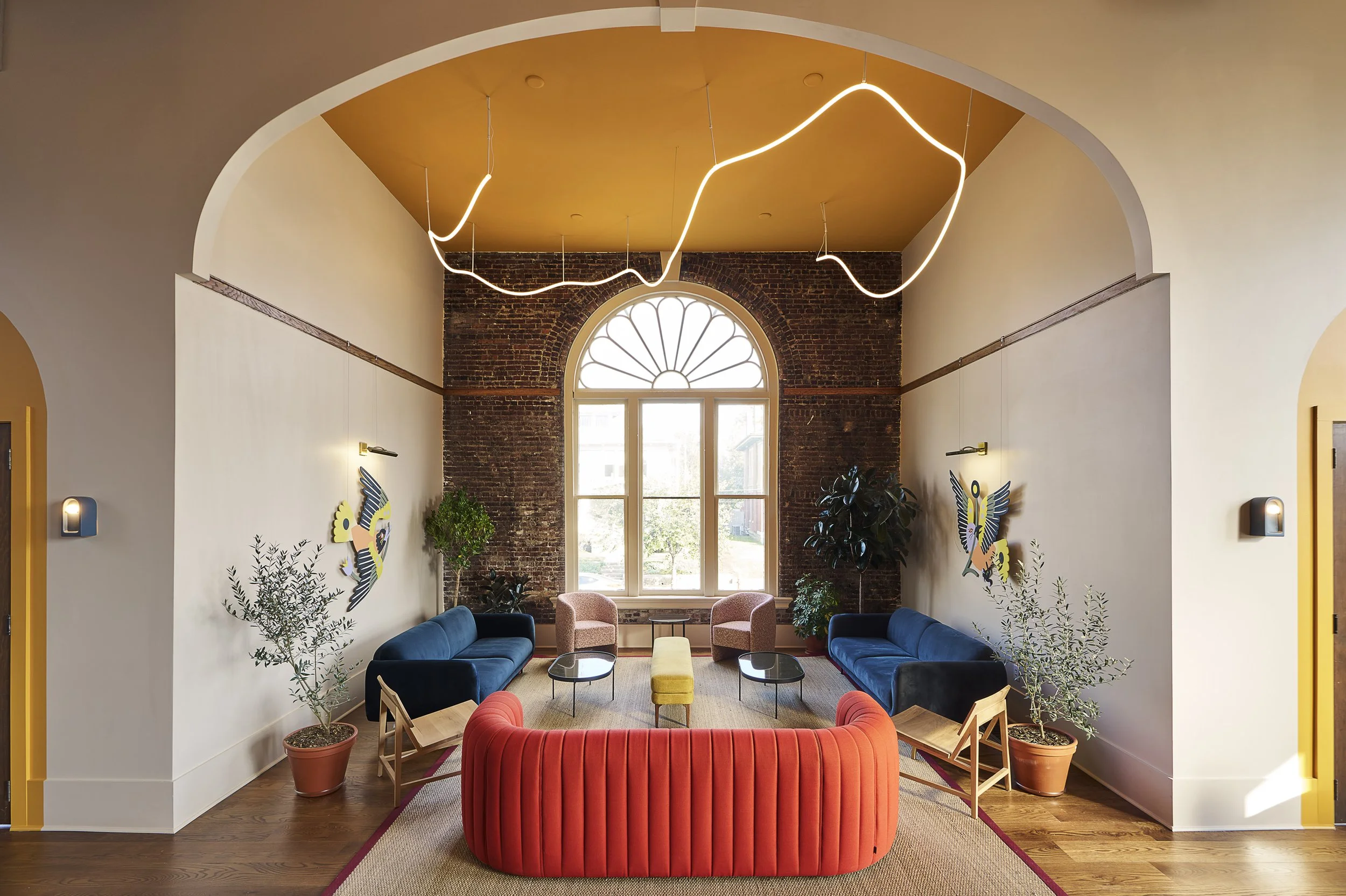 A modern and cozy living room with a exposed brick wall and large arched window. There is a yellow ceiling with a modern wavy light fixture. The room features two blue velvet sofas, two pink armchairs, and various potted plants. The space is decorate