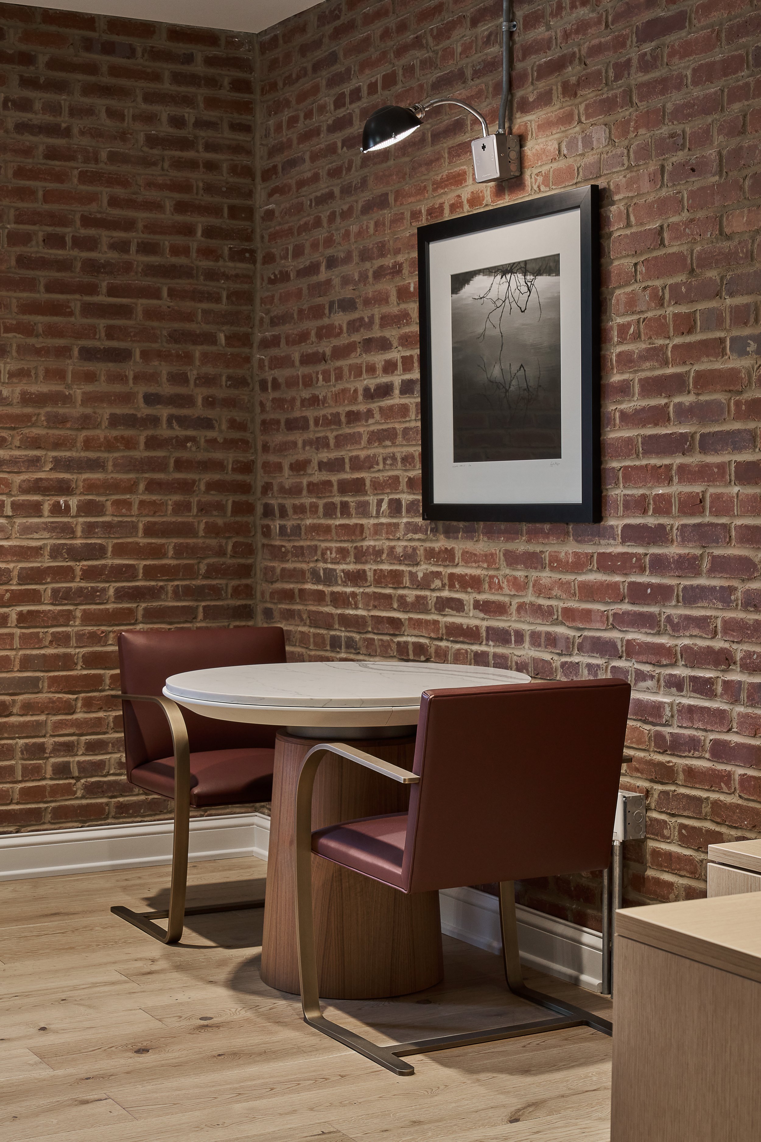 A cozy dining area with two wooden chairs with muted red cushions around a small, white, round table. The background features a red brick wall with a framed black-and-white photograph of a tree reflected in water. An adjustable wall lamp with a black