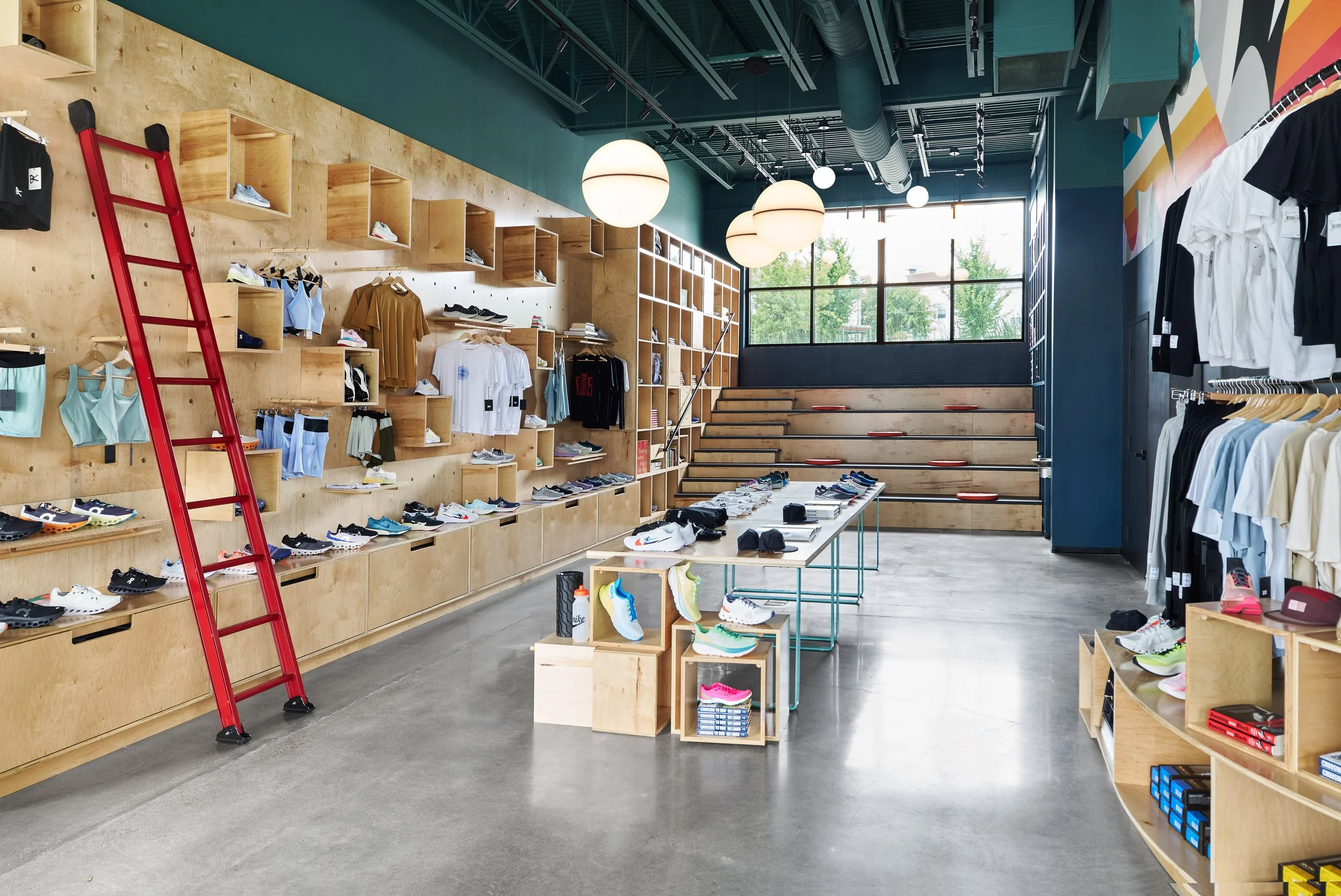 Inside a modern retail sneaker store with wooden wall displays, shelves, and tables showcasing athletic shoes; retail ladder leaning against the wall, large window letting in natural light, and colorful sneakers arranged on tables.