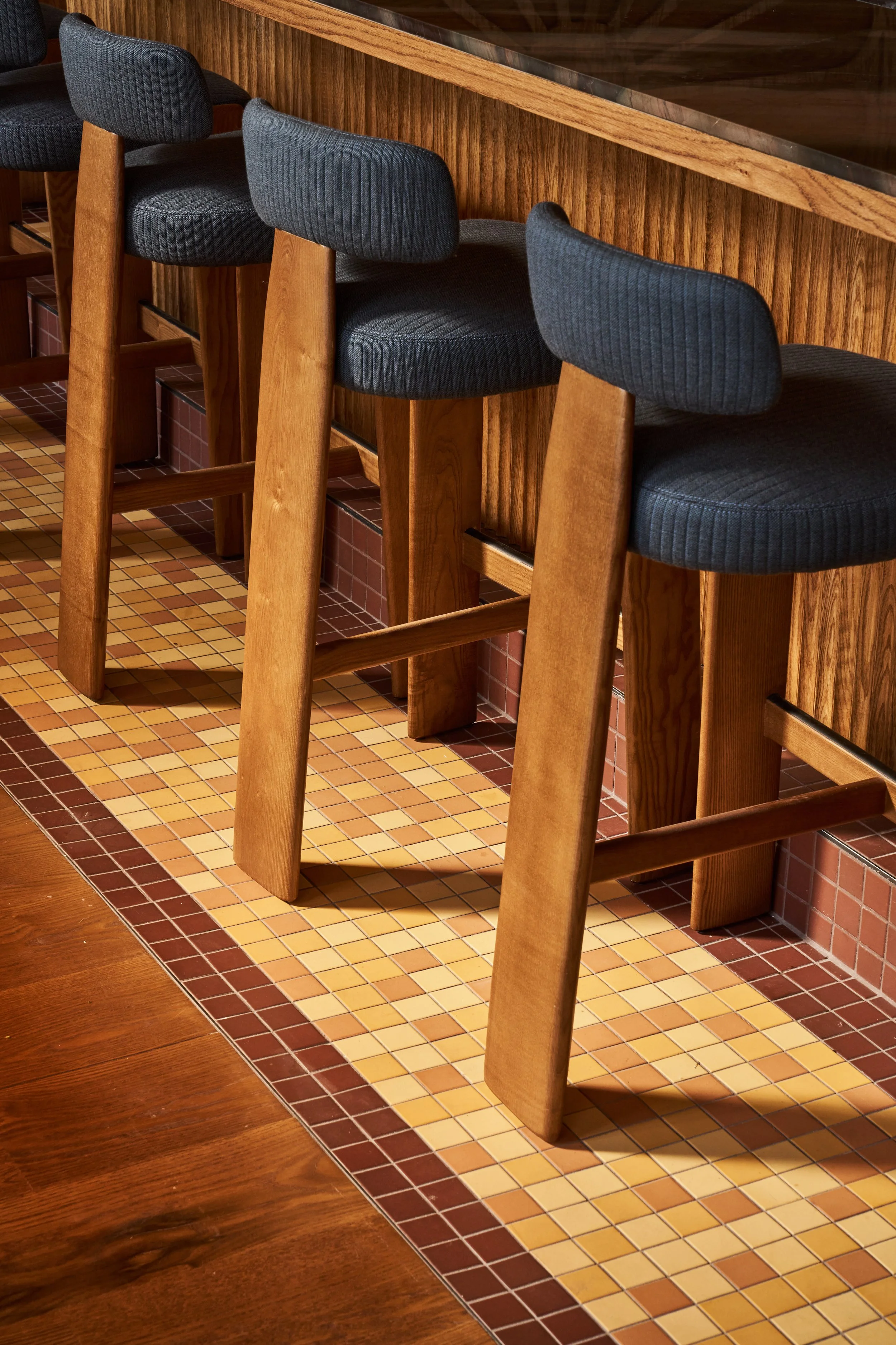 Wooden chairs with dark gray upholstered seats and backs lined up against a wooden bar counter in a restaurant or cafe with tiled flooring.