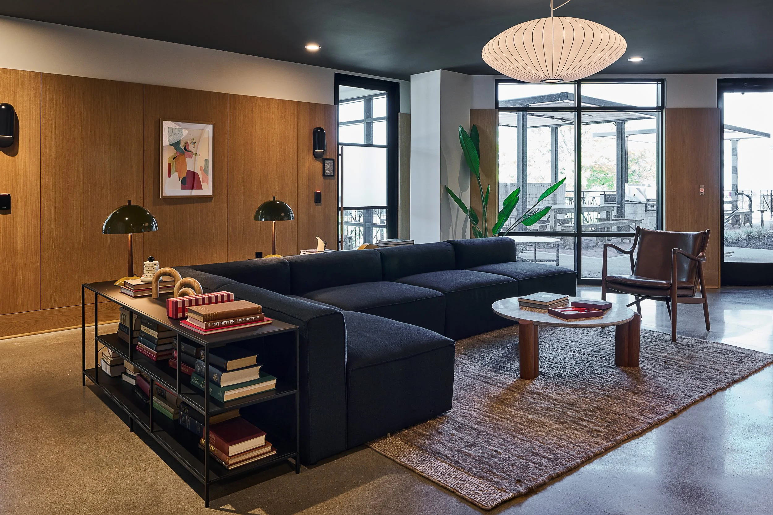 Modern interior living room with a black sofa, a coffee table, a bookshelf filled with books, a large rug, wooden walls, plants, pendant lighting, and large windows showing outdoor seating.