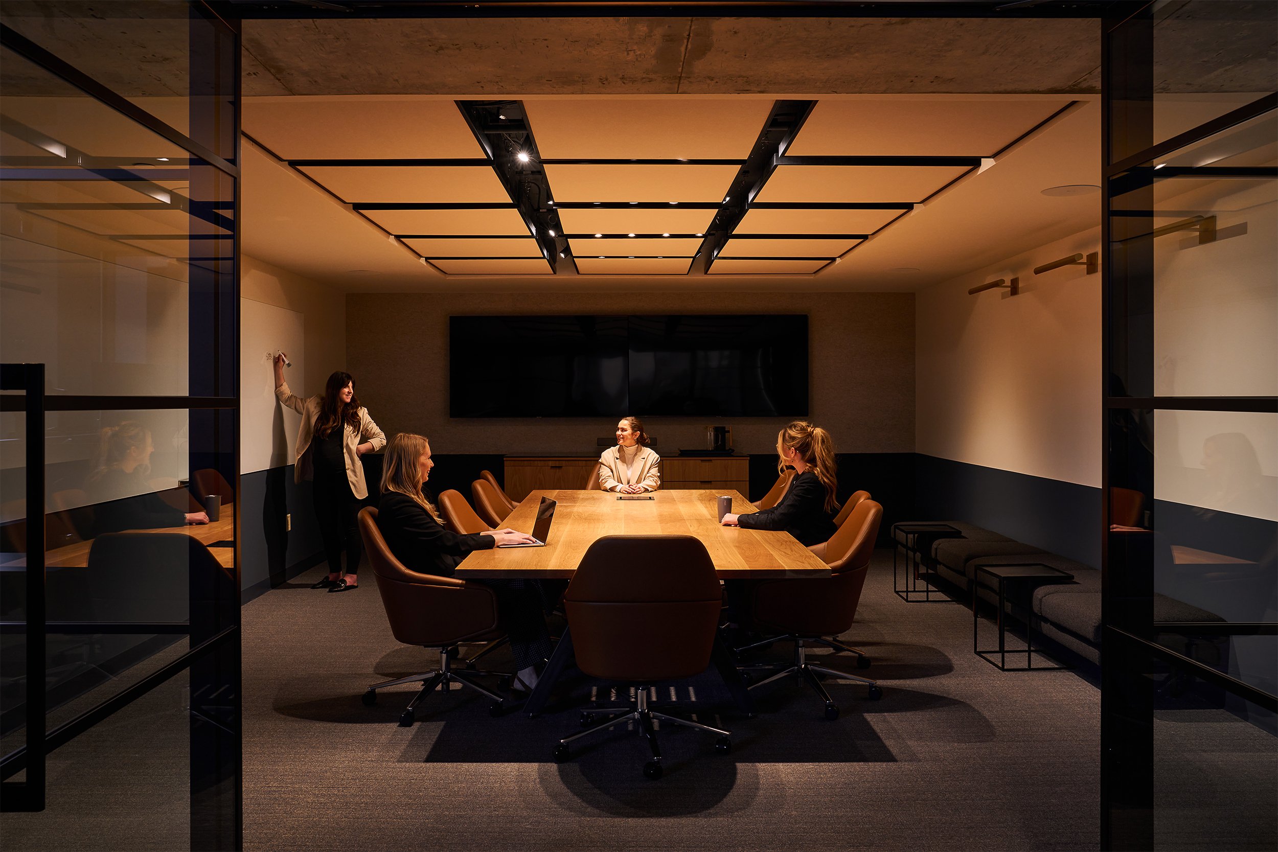 A modern conference room with a long wooden table, several women sitting and a woman standing and speaking at a whiteboard, with large screens on the wall and glass walls on either side.
